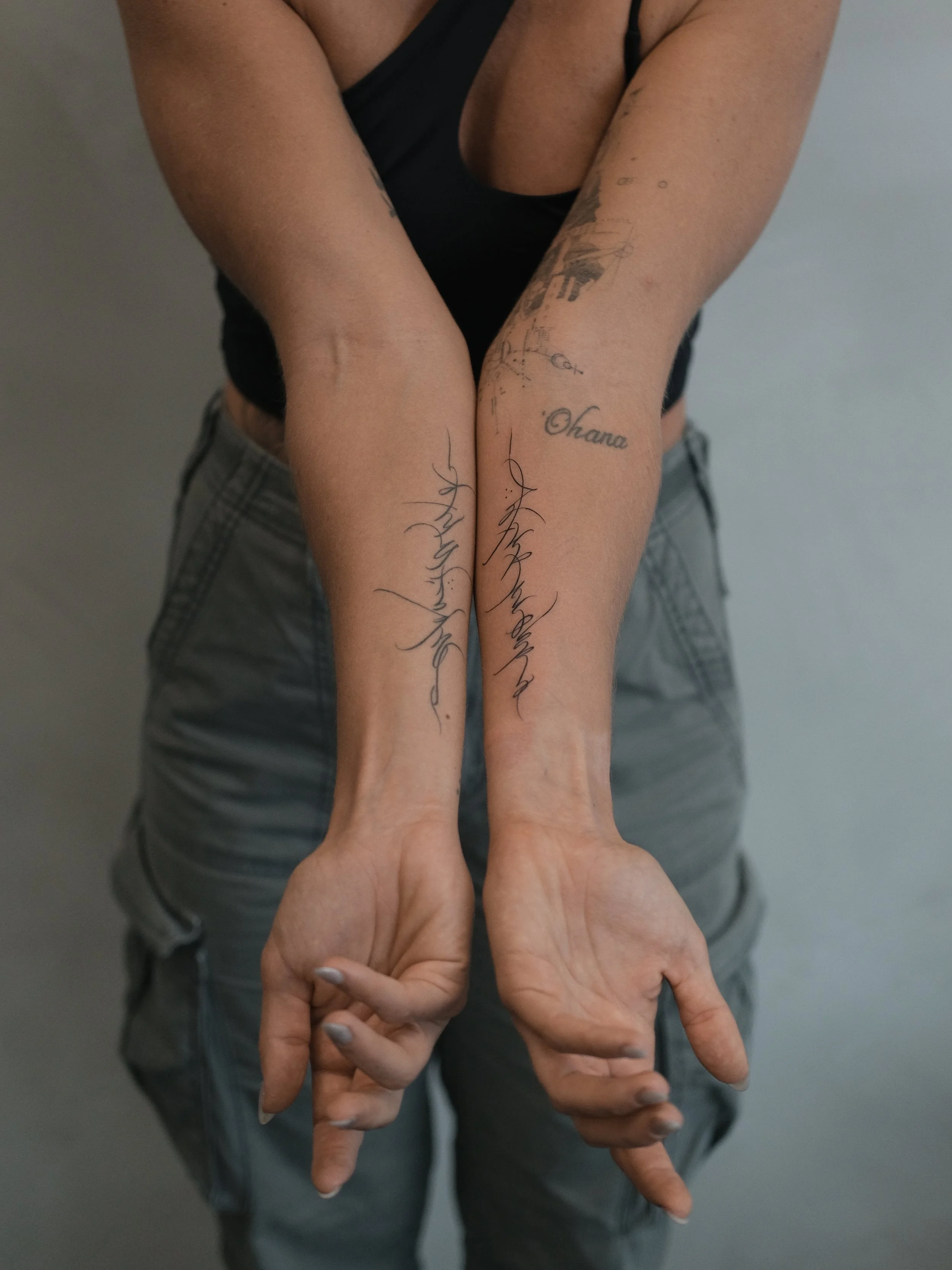 A person is seen from the waist up, with their arms extended downward and hands crossed at the wrist in front of them. They are wearing a black armless top and gray plaid shorts. The person has tattoos on their forearms, including the words 'Ohana' a