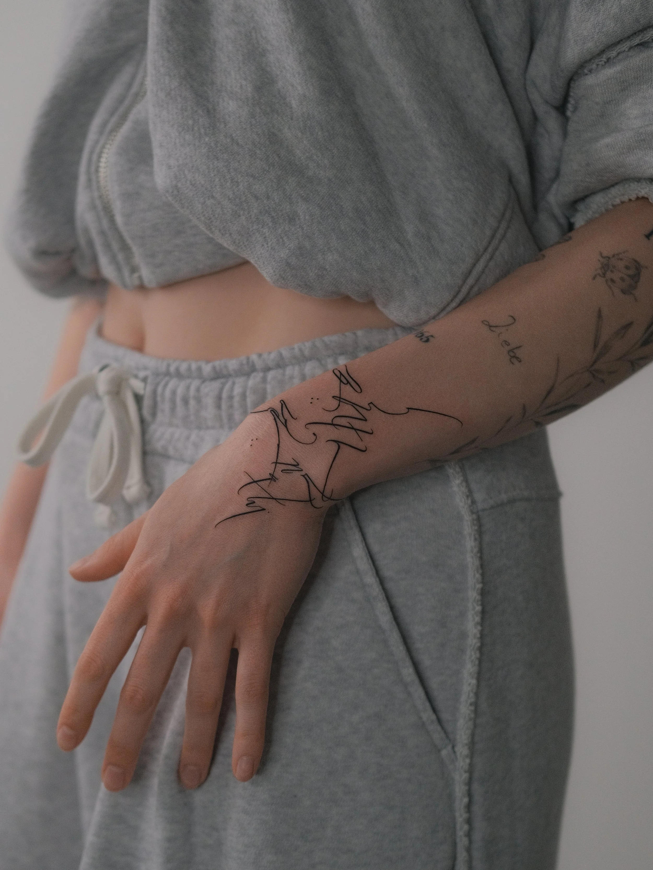 Close-up of a person's arm with a tattoo, wearing grey sweatpants and a grey hoodie.