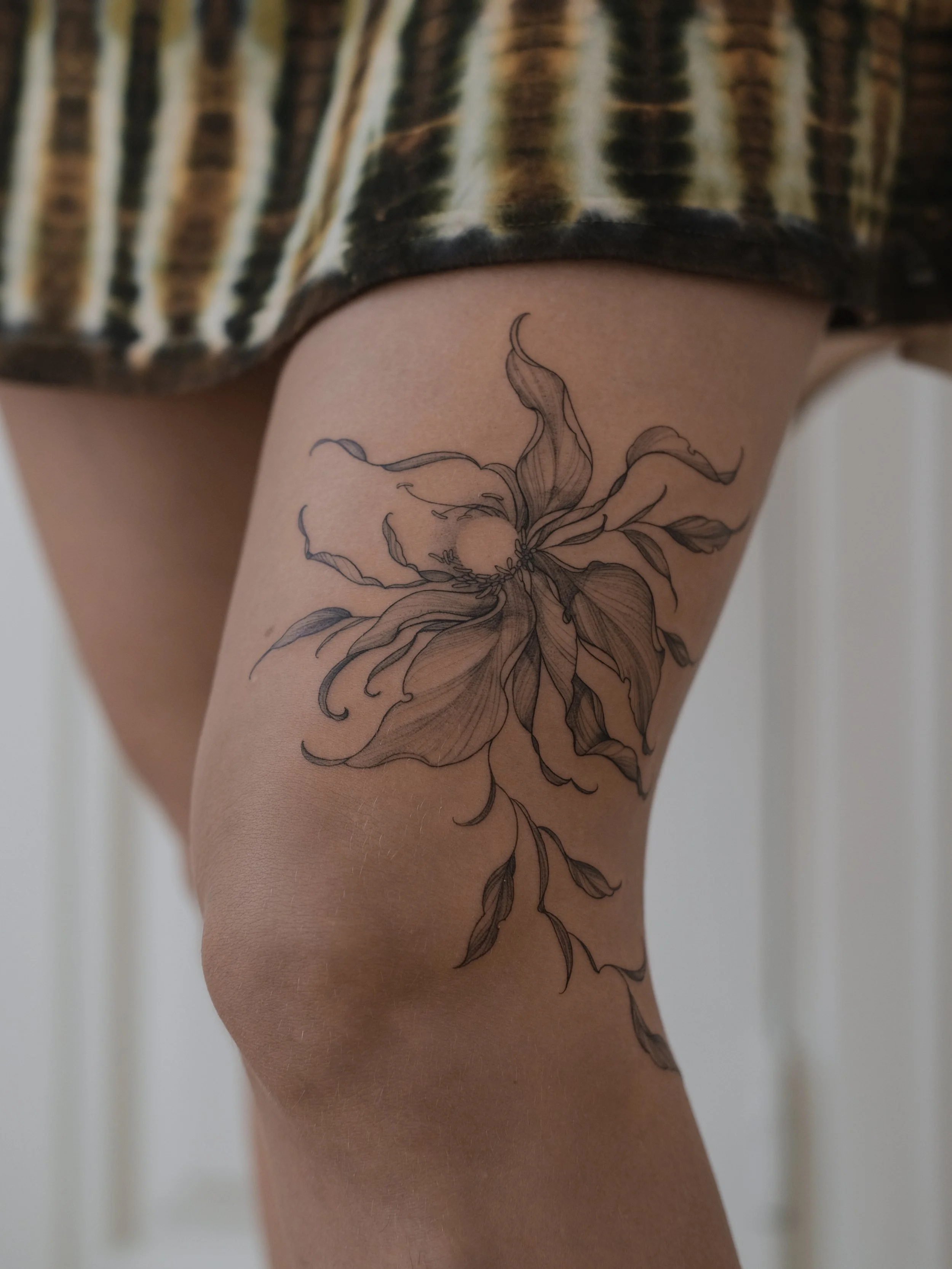Close-up of a person's thigh with a black outline tattoo of a detailed flower with flowing petals and leaves, wearing a striped skirt.