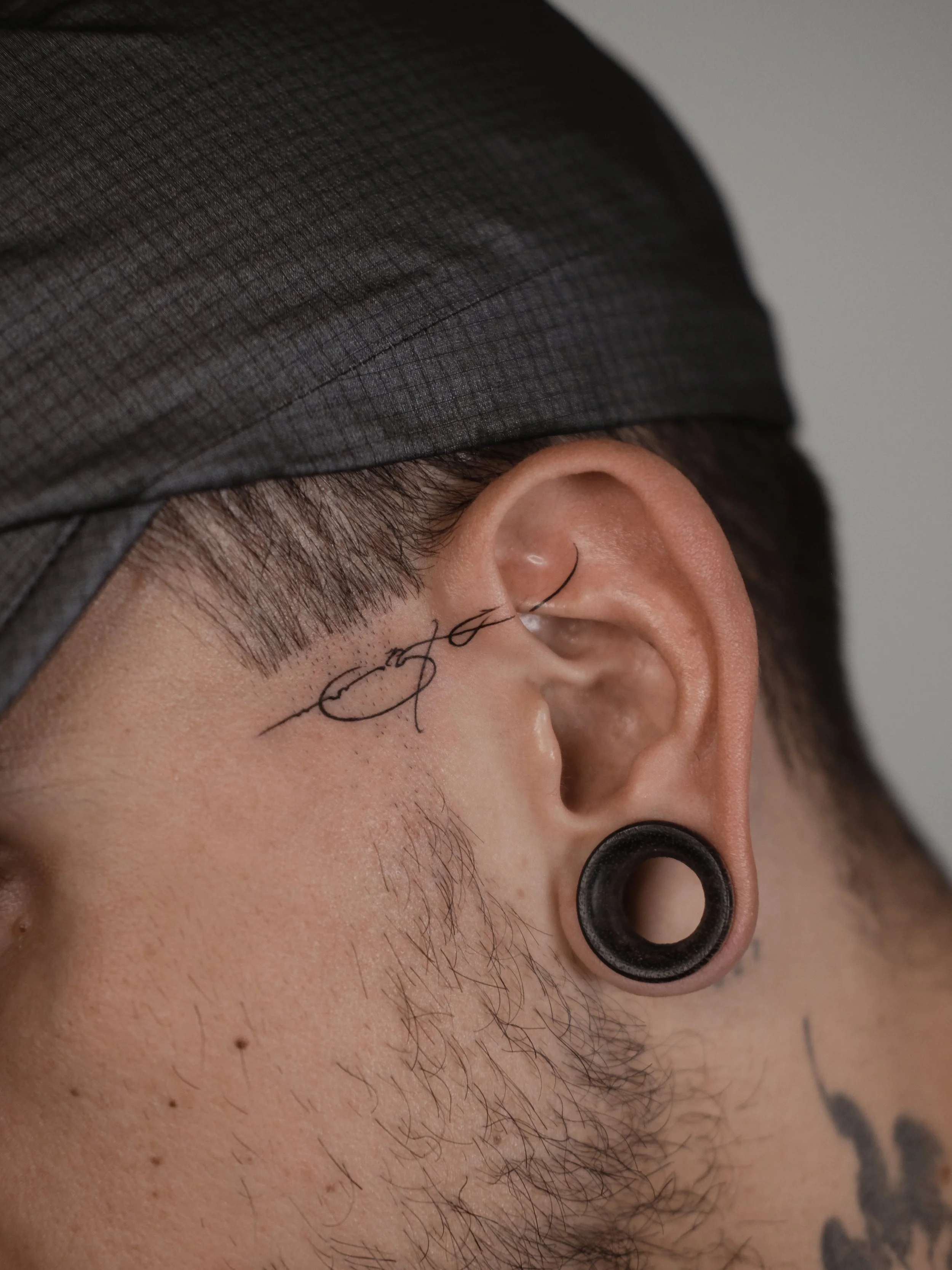 Close-up of a person's ear with black gauges, a tattoo behind the ear, and a partial view of a face wearing a dark gray cap.