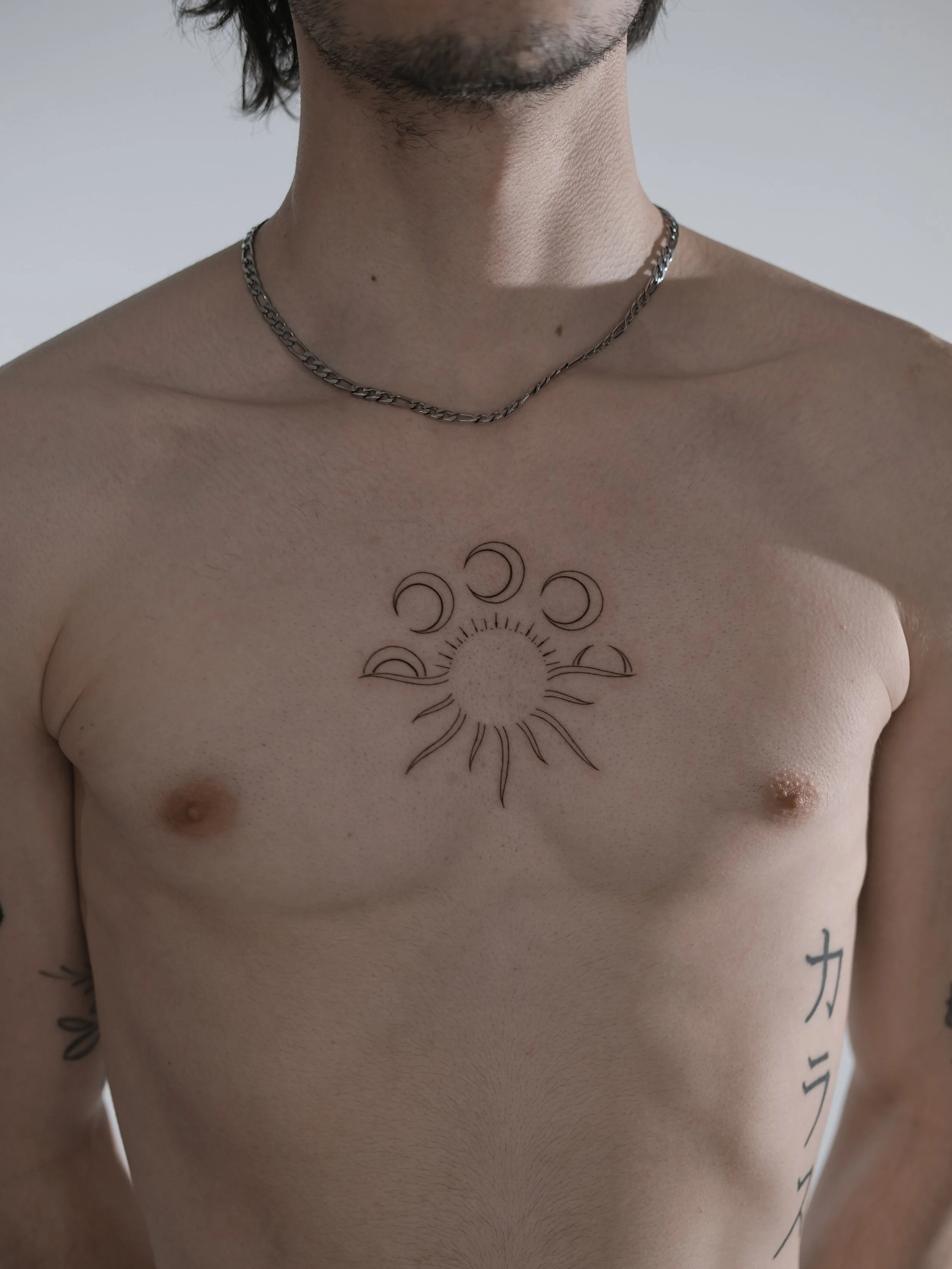 A shirtless person with a tattoo of a sun with crescent moon phases on the chest, wearing a chain necklace, with visible facial hair and partial shoulder tattoos.