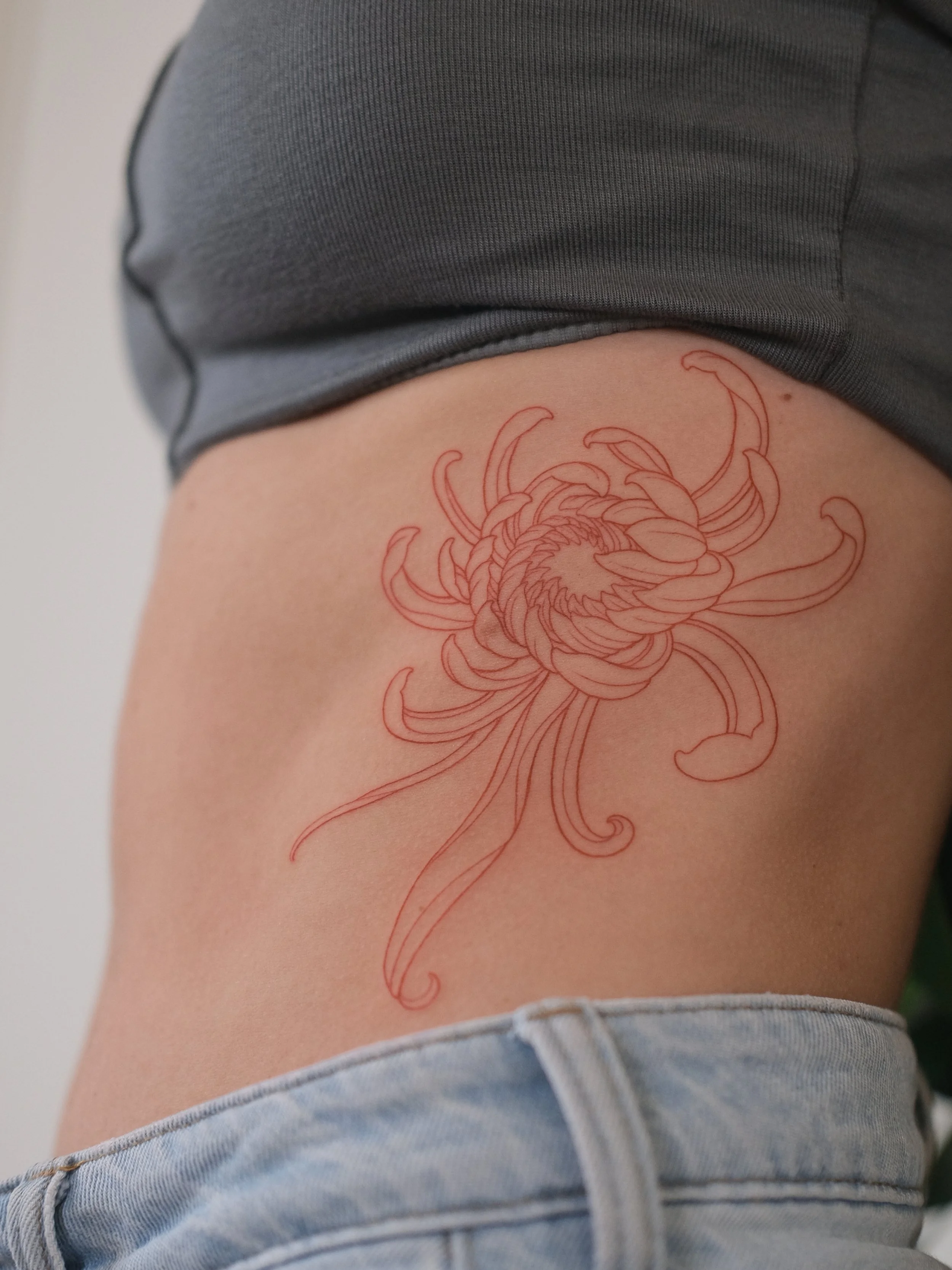 A person's side torso with a red ink outline of a flower tattoo on the upper thigh, wearing denim shorts and a dark gray top.