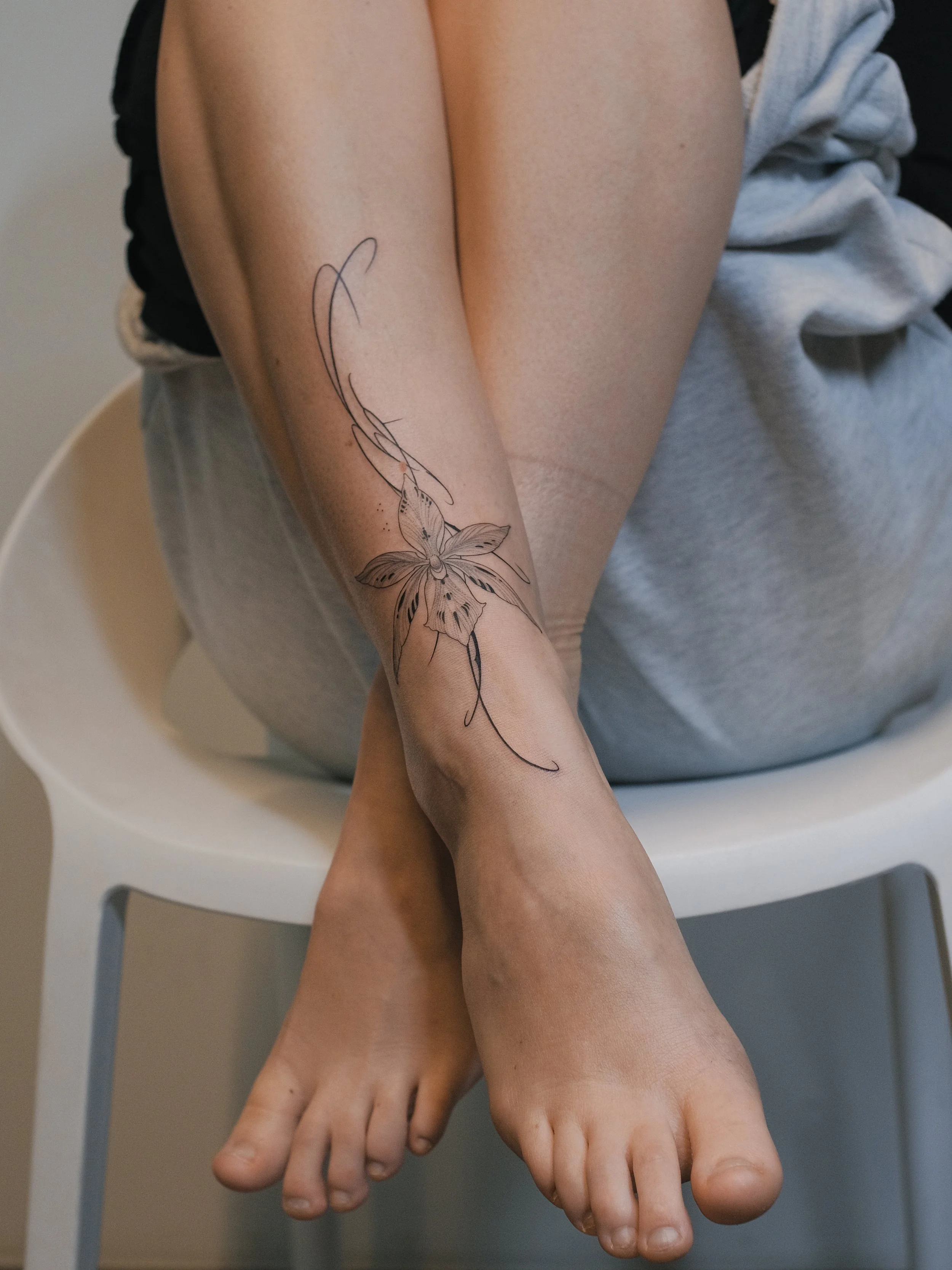 A person's legs and feet with a tattoo of a butterfly on the lower leg, seated on a white chair. The person is wearing gray shorts and has a blanket or clothing draped over their lap.