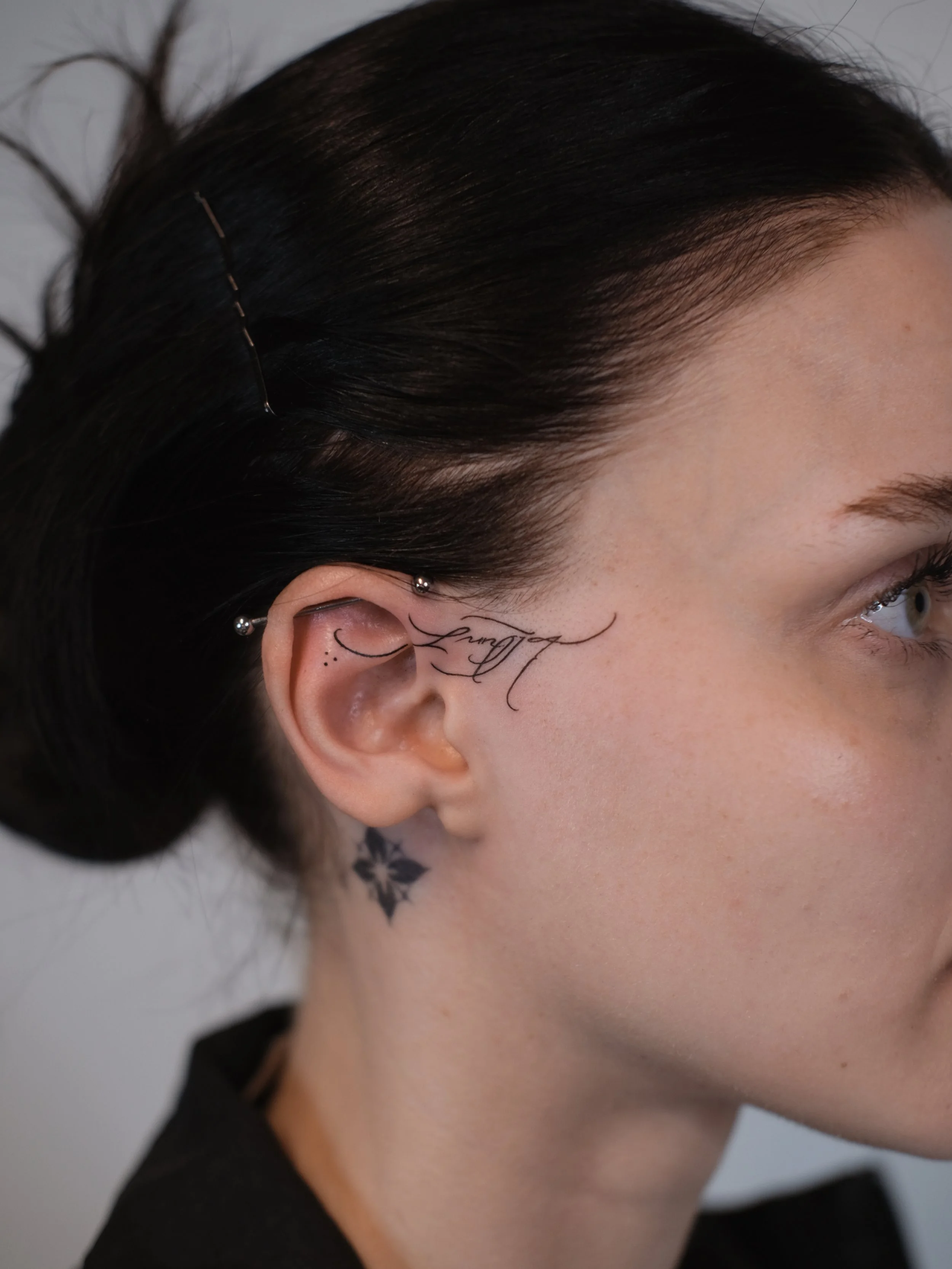 Close-up of a person's right side face showing black hair with hairpins, eye, and a tattoo on the neck.