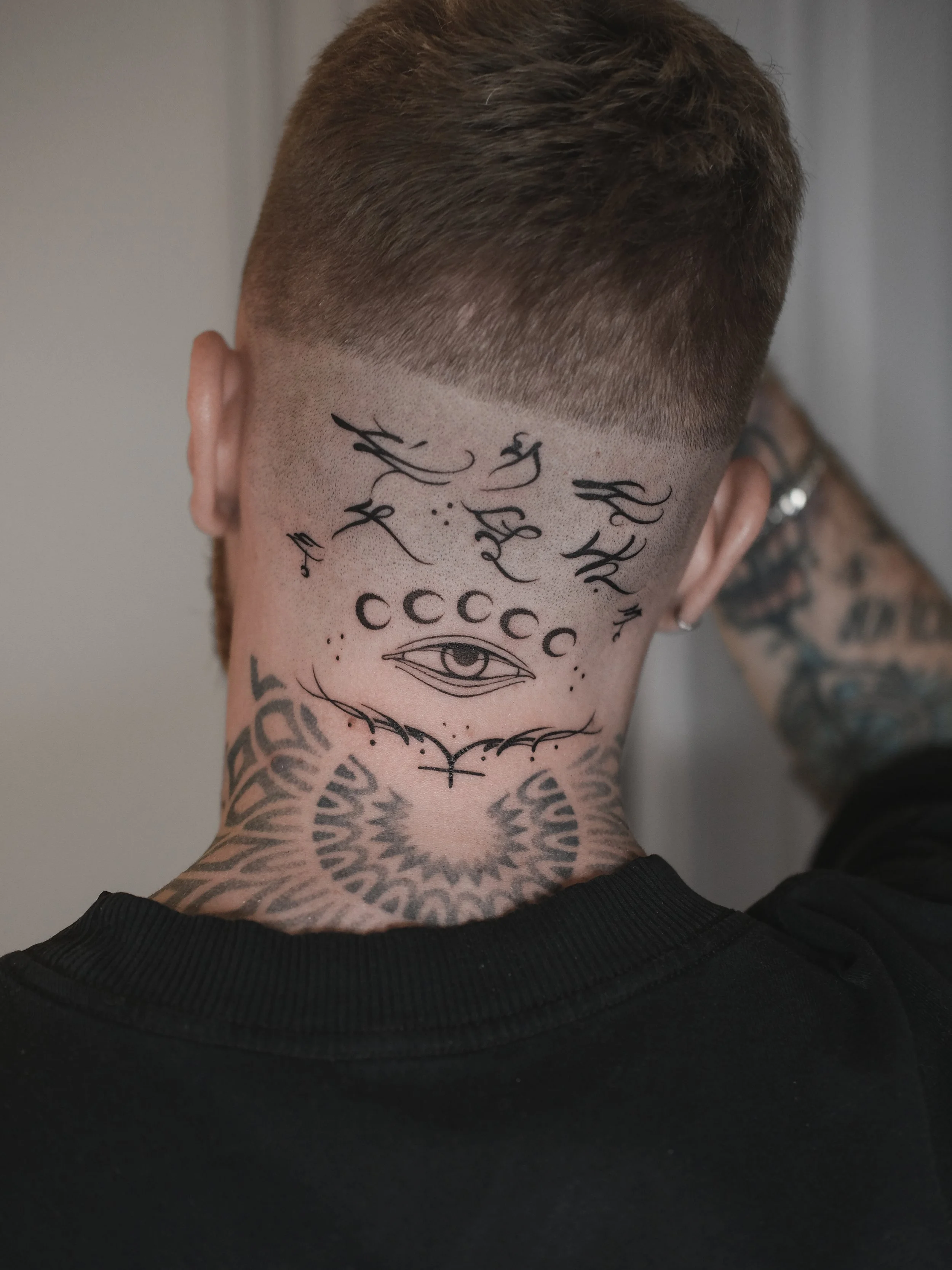 Back of a person's head and neck showing a detailed tattoo design featuring an eye, moon phases, and intricate patterns, with a fade haircut.