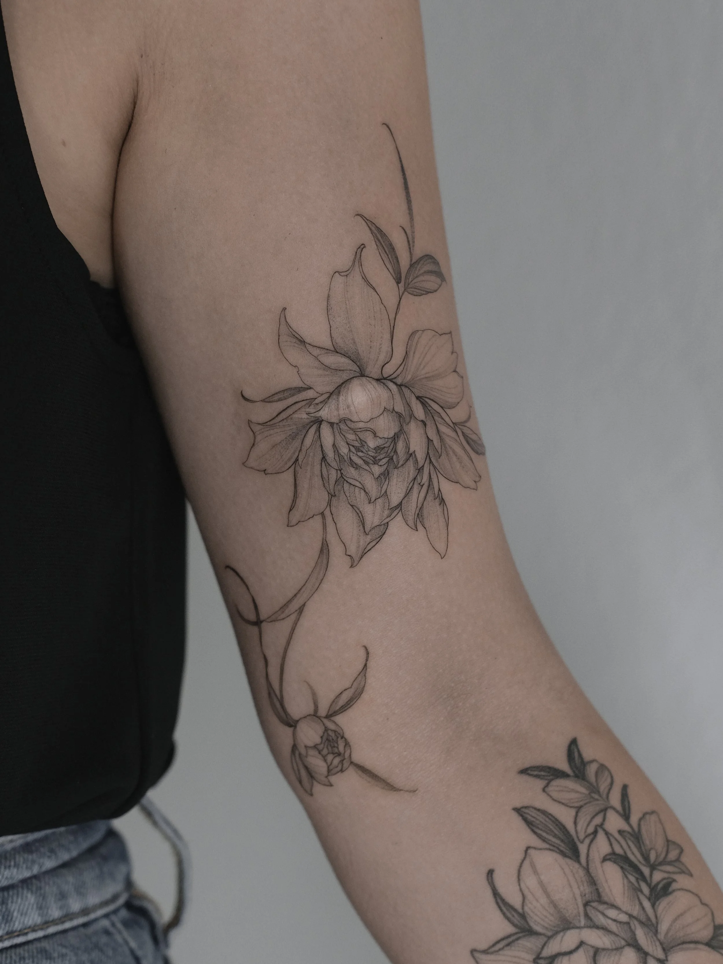 A person's arm with a detailed black and gray floral tattoo that includes flowers, leaves, and stems.