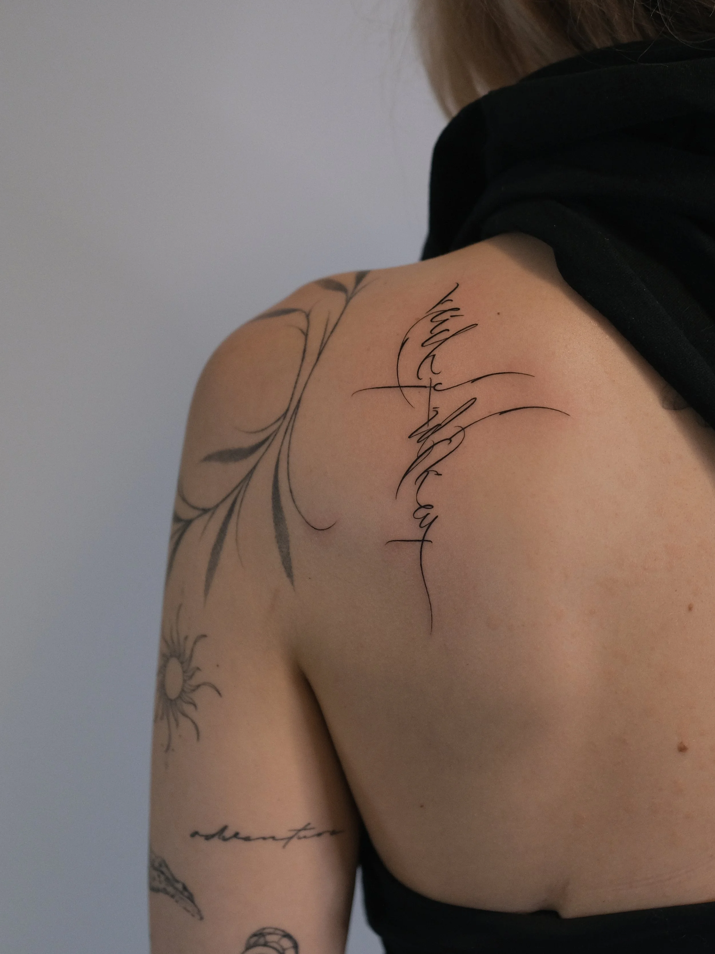 Close-up of a person's shoulder and upper arm revealing tattoos with cursive writing and floral designs on light skin and wearing a black top.