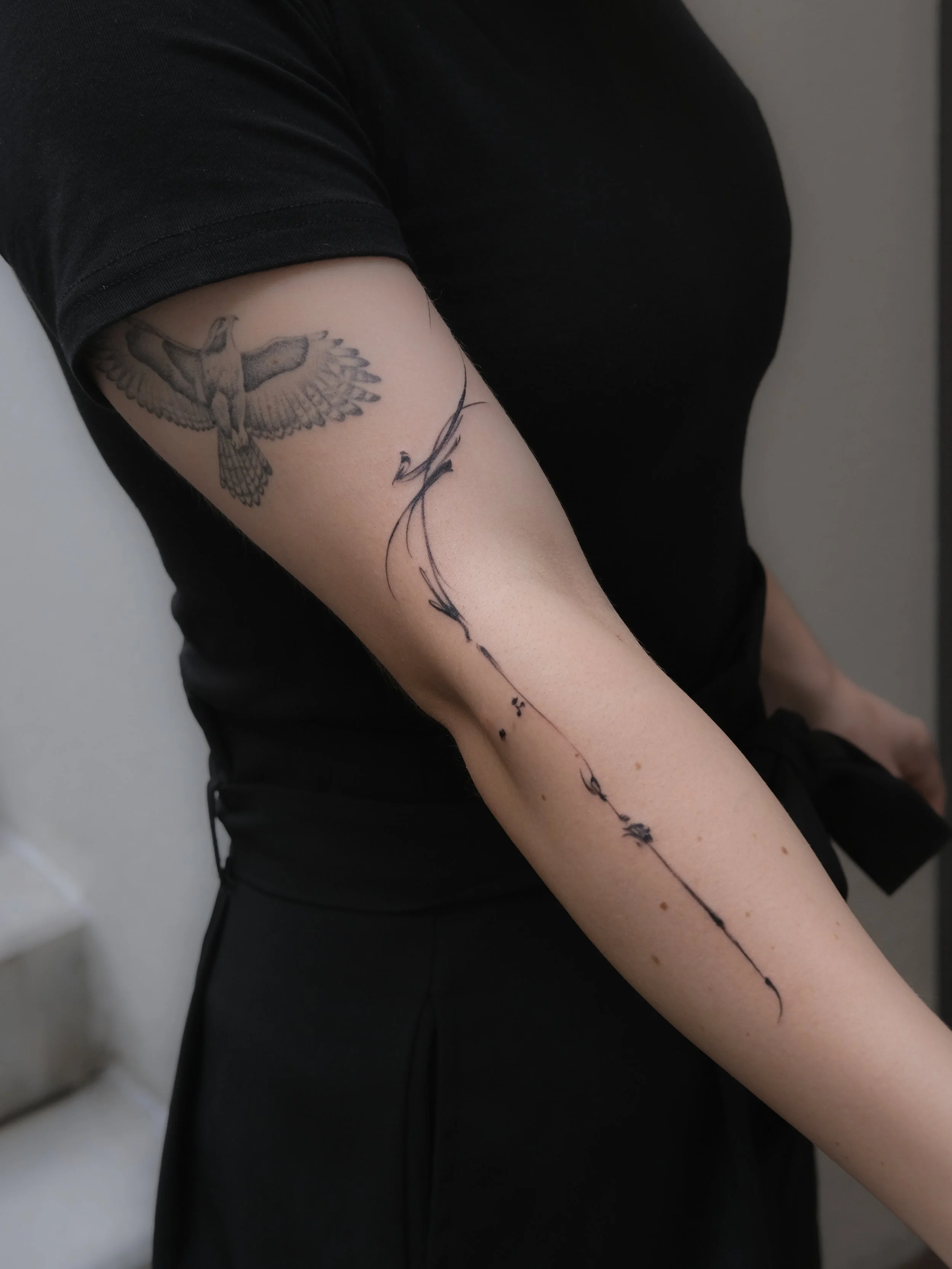Close-up of a person's arm with a black and gray bird tattoo on the upper arm and a delicate black script tattoo running down the forearm, wearing a black short-sleeve shirt.