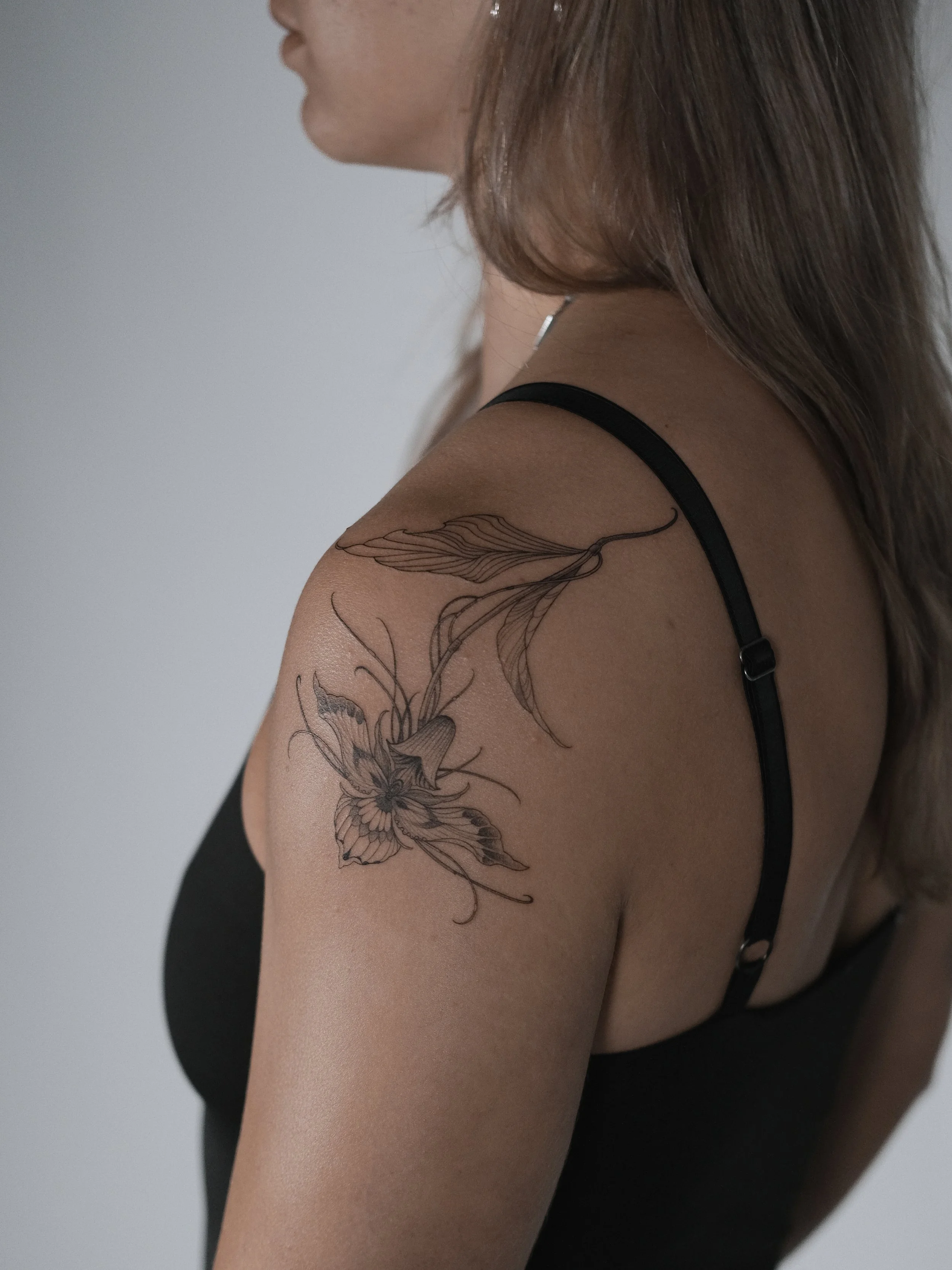 A woman with brown hair wearing a black spaghetti strap top. She has a floral and butterfly tattoo on her upper arm.