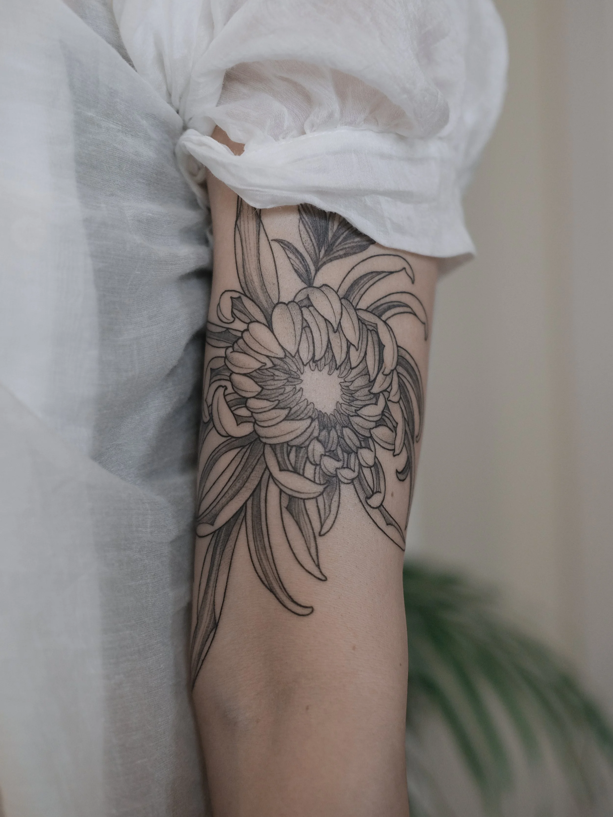 A person's arm with a large black outline floral tattoo visible near the elbow, partially covered by a white, billowy sleeve or fabric.