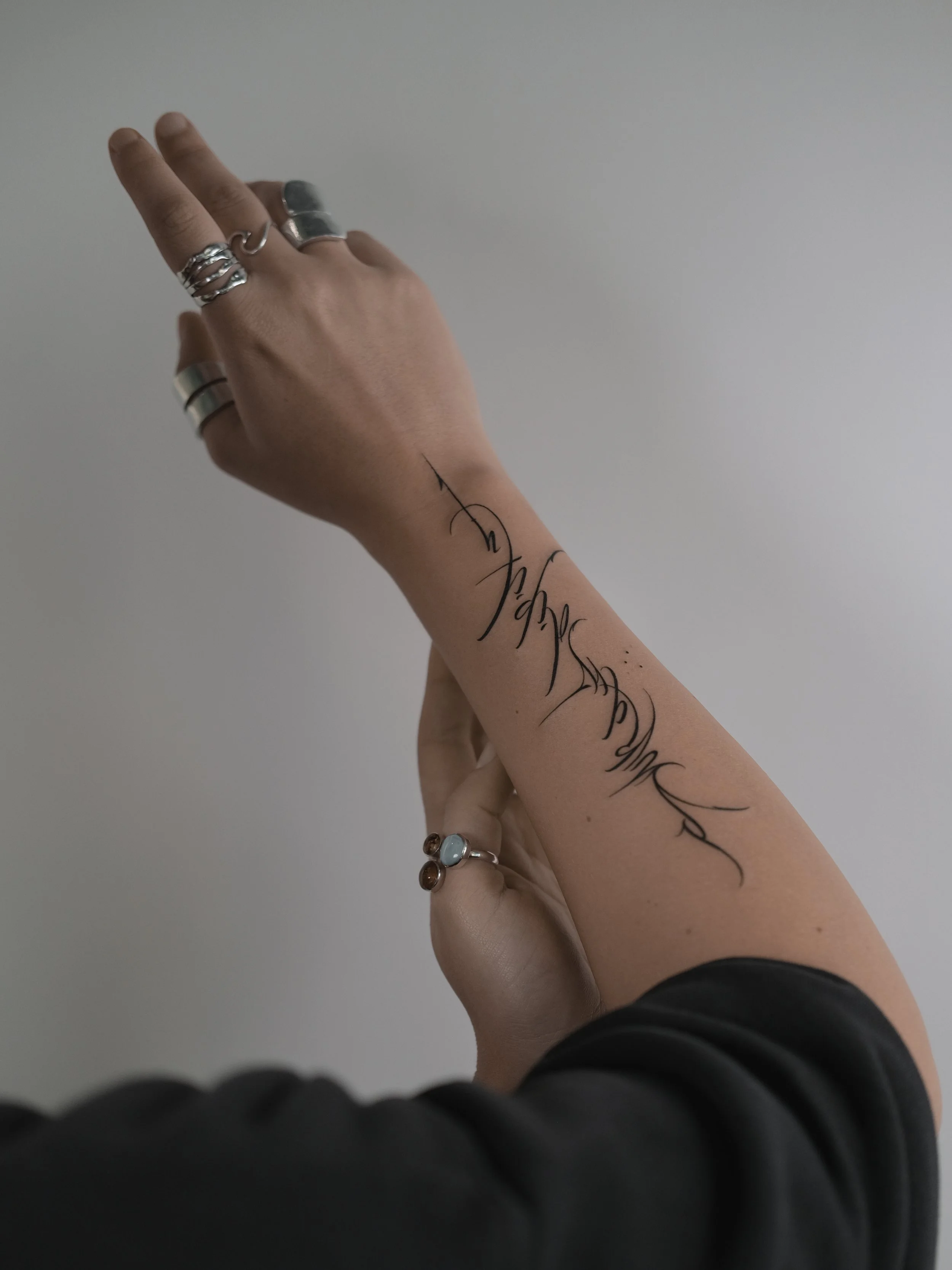 A person's hand and wrist with multiple rings, with a tattoo on their forearm that reads 'dream'. The background is plain, and the person is wearing a black long-sleeve shirt.