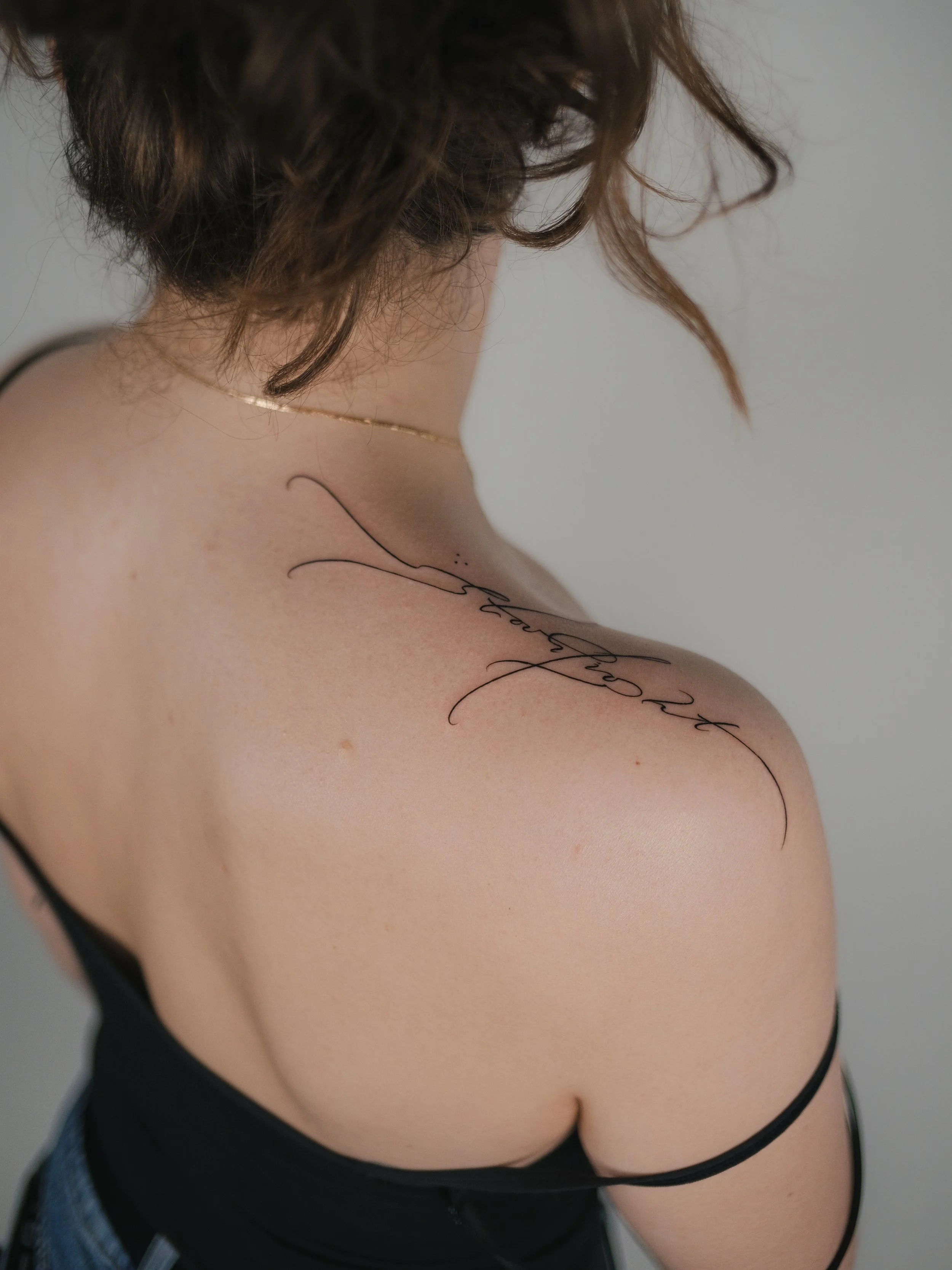 Partial side view of a woman with curly brown hair wearing a black tank top and jeans, with a tattoo on her shoulder.