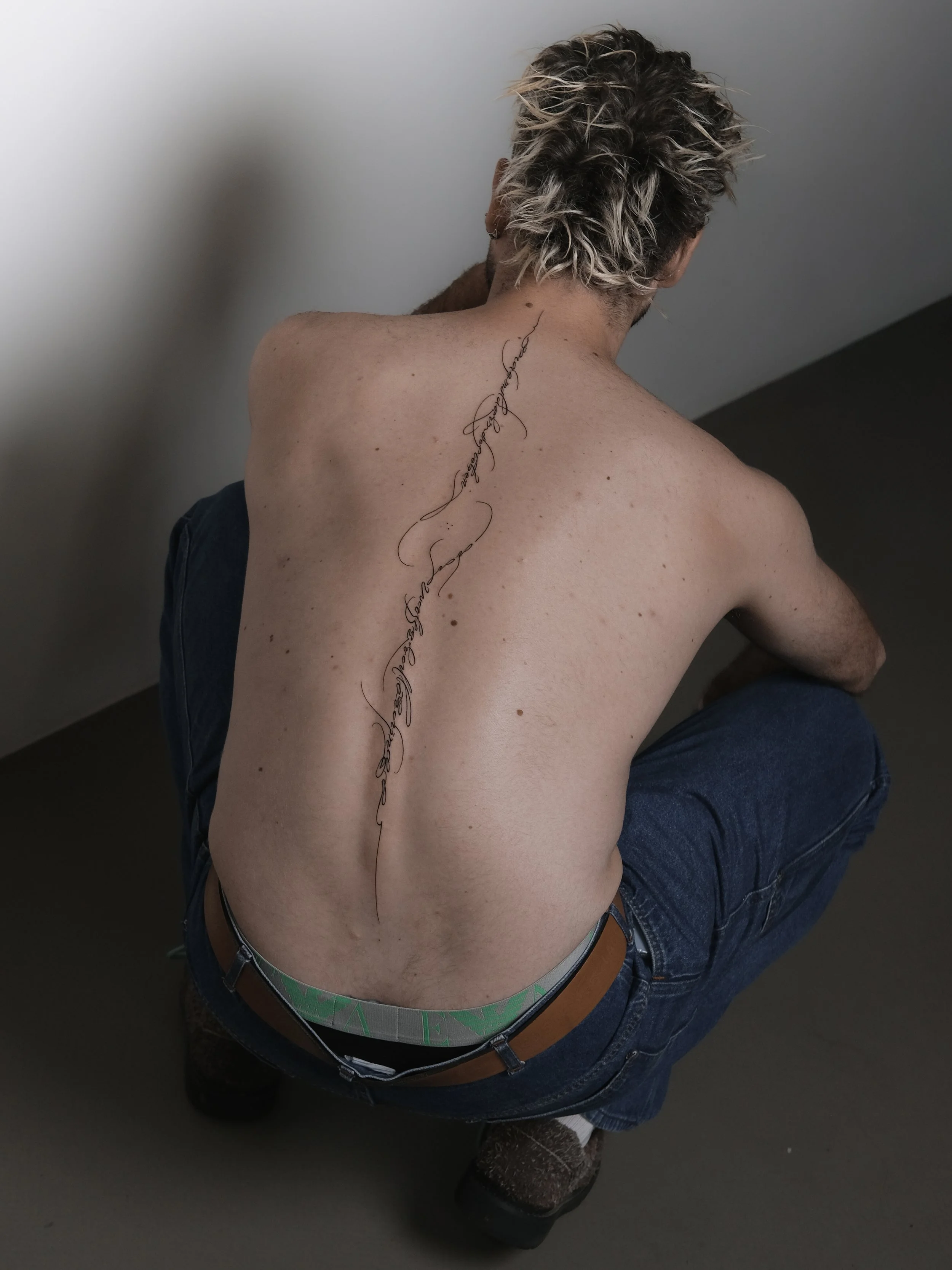 A shirtless man with curly, light-colored hair sitting on the floor, with a tattoo running down the back of his neck.