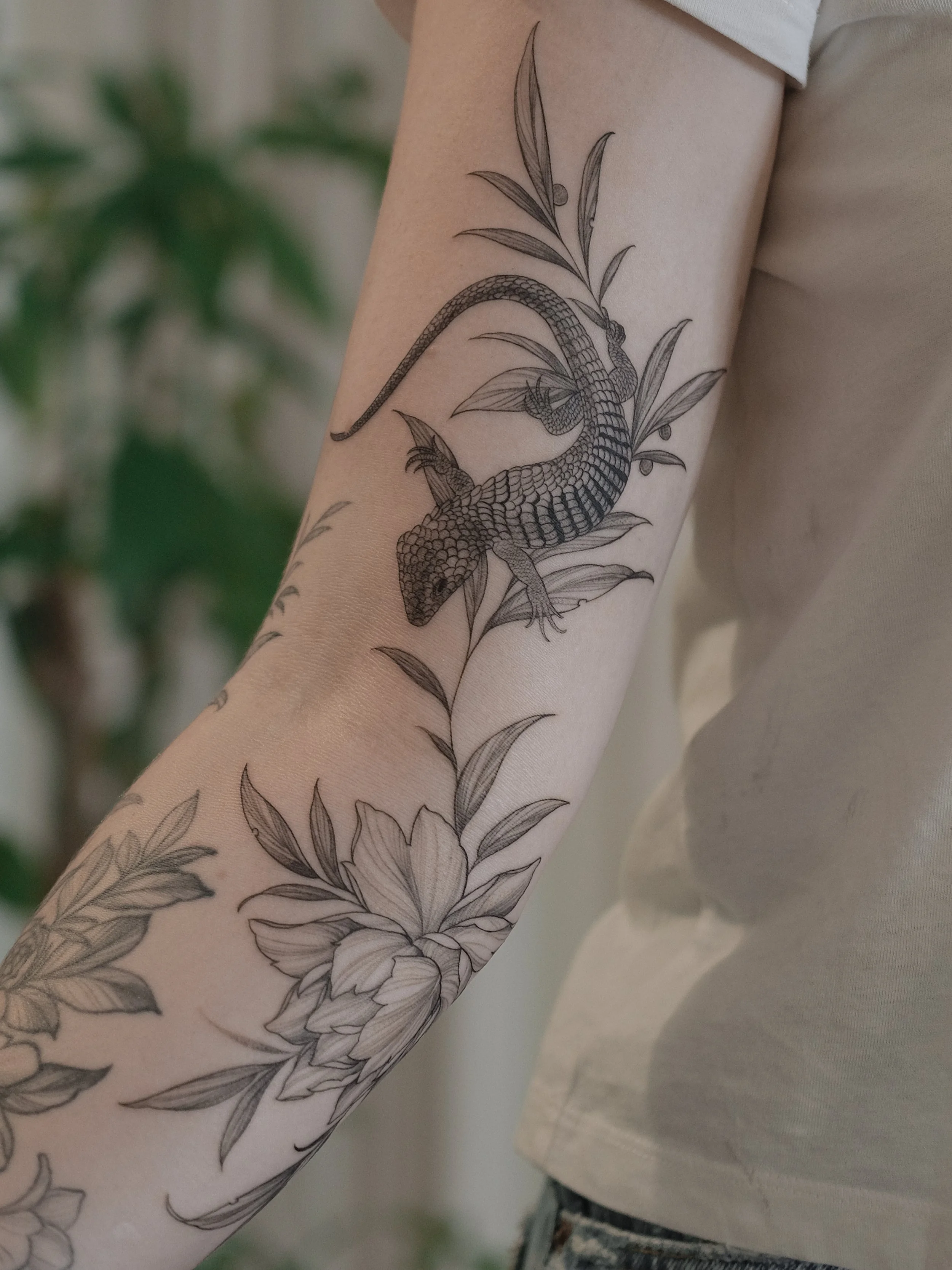 A detailed black and gray tattoo of a snake intertwined with leaves and flowers, covering most of an arm.