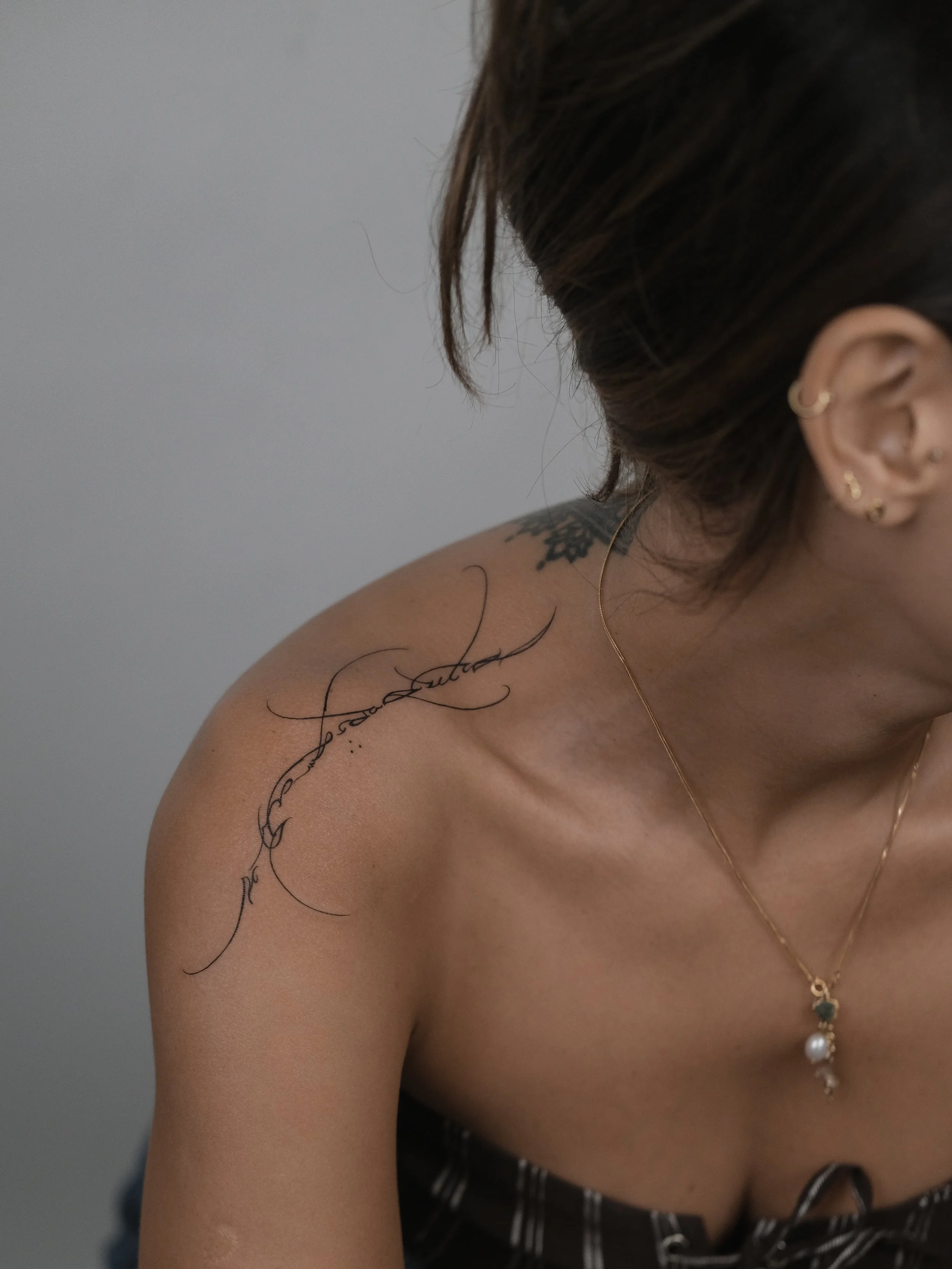A woman with dark hair and multiple earrings is showing her left shoulder which has a tattoo and is adorned with jewelry, including a necklace with pendants.