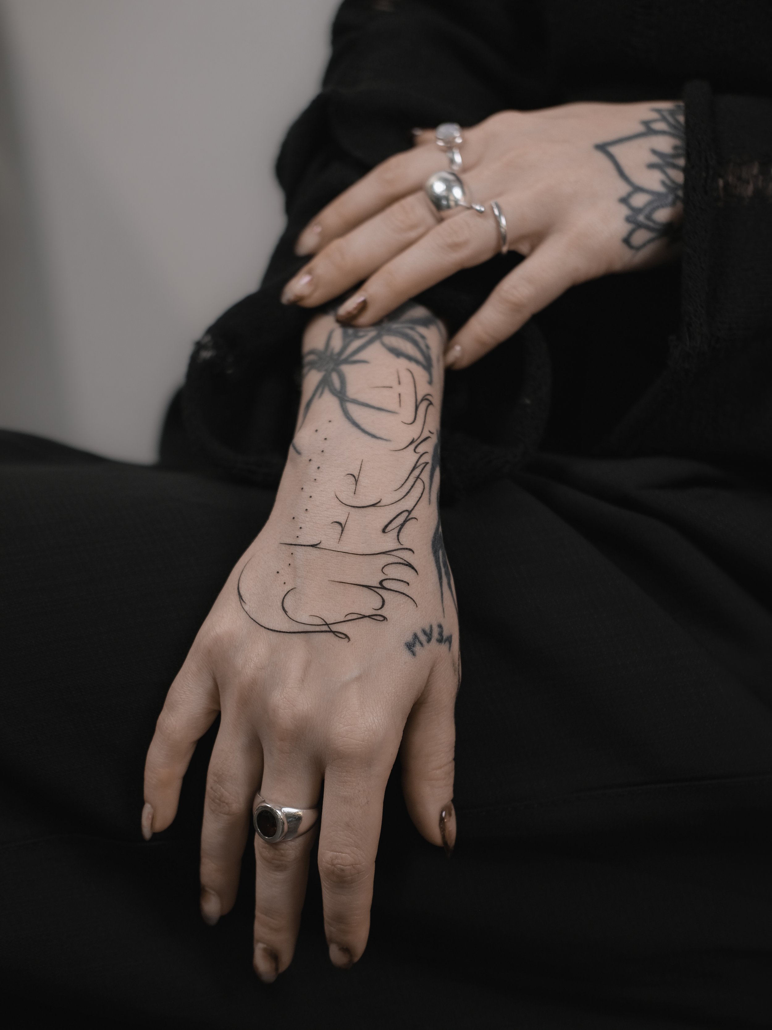 A person's hand resting on their lap, adorned with multiple rings and tattoos. The tattoo on the wrist features black ink script and a floral design, while the person's nails are painted a dark color, and a silver ring with a large stone is worn on t