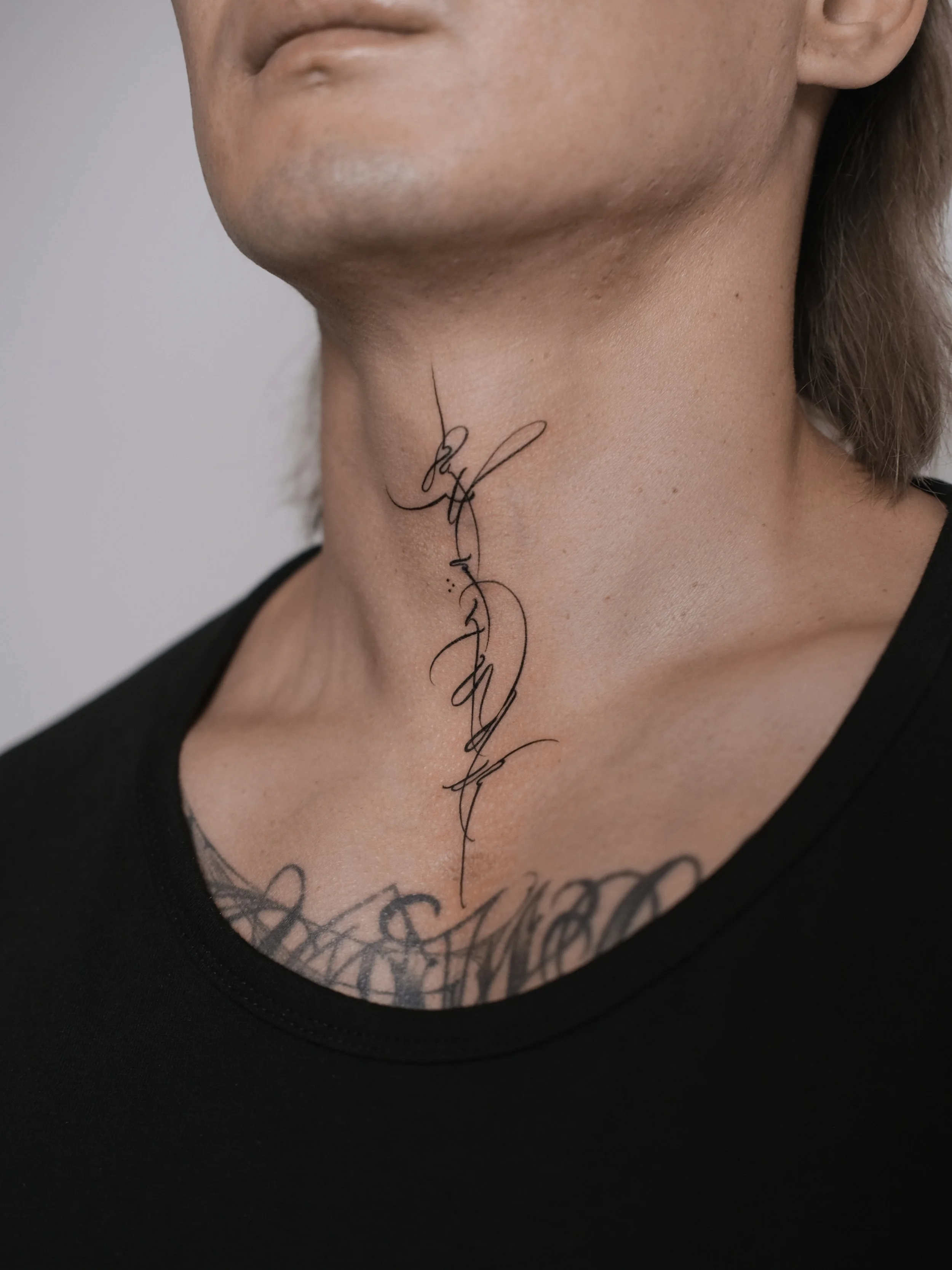 Close-up of a person's neck with a black tattooed script design and a tattooed design visible underneath the black shirt.