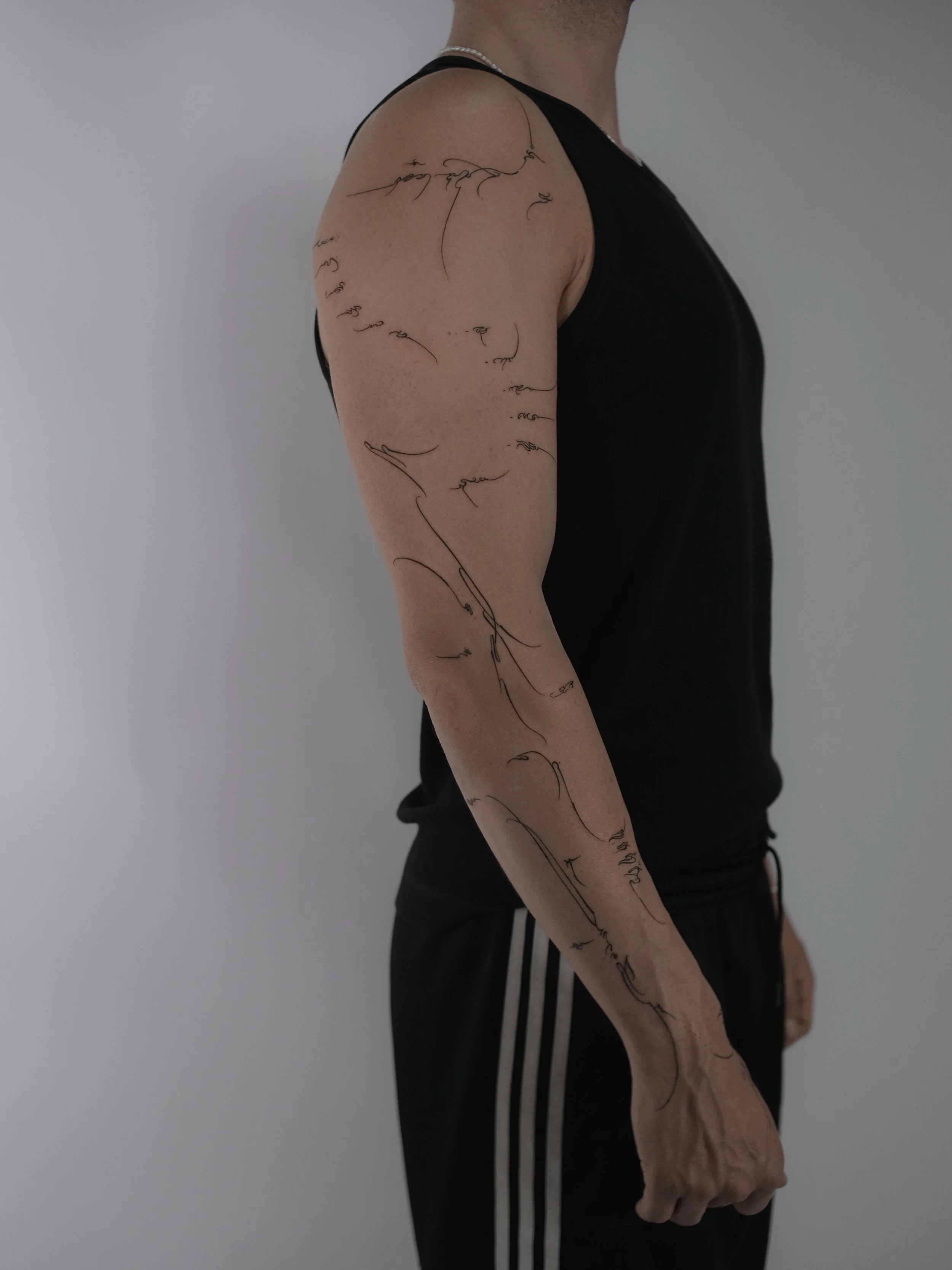 A person's left arm with a tattoo of a continuous line drawing of a face and a neck, seen in profile, extending from shoulder to wrist, wearing a black sleeveless shirt and black athletic pants with white stripes.