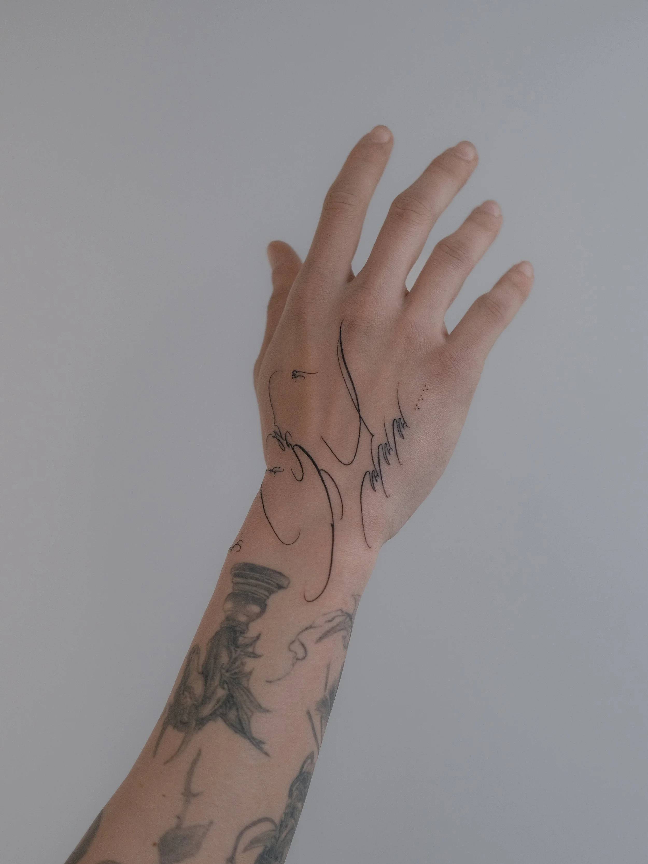 A hand with multiple tattoos stretching upward, with a plain, light-colored background.