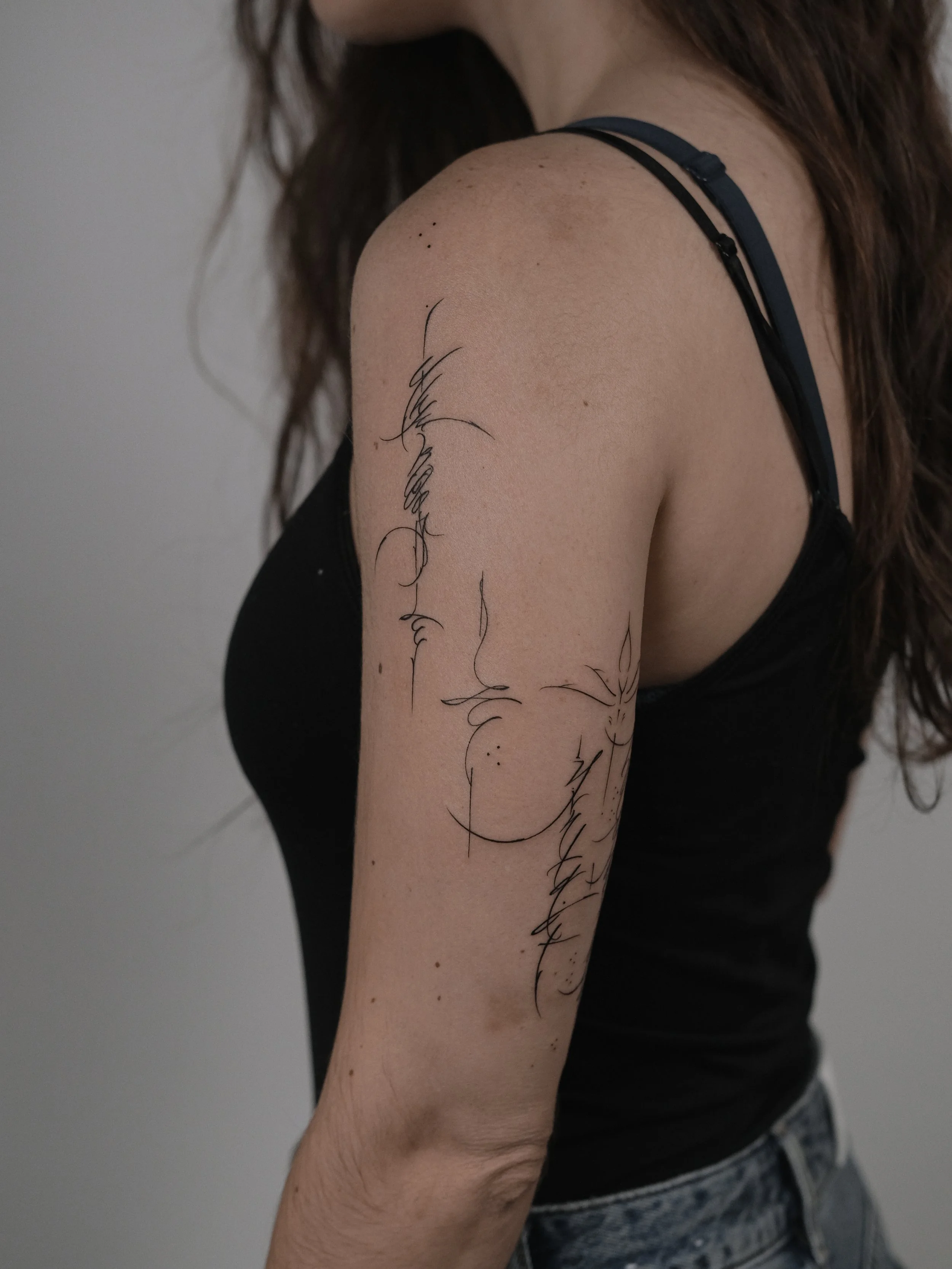 Close-up of a woman's arm with black line art tattoos of faces and text, wearing a black spaghetti strap top and blue jeans.