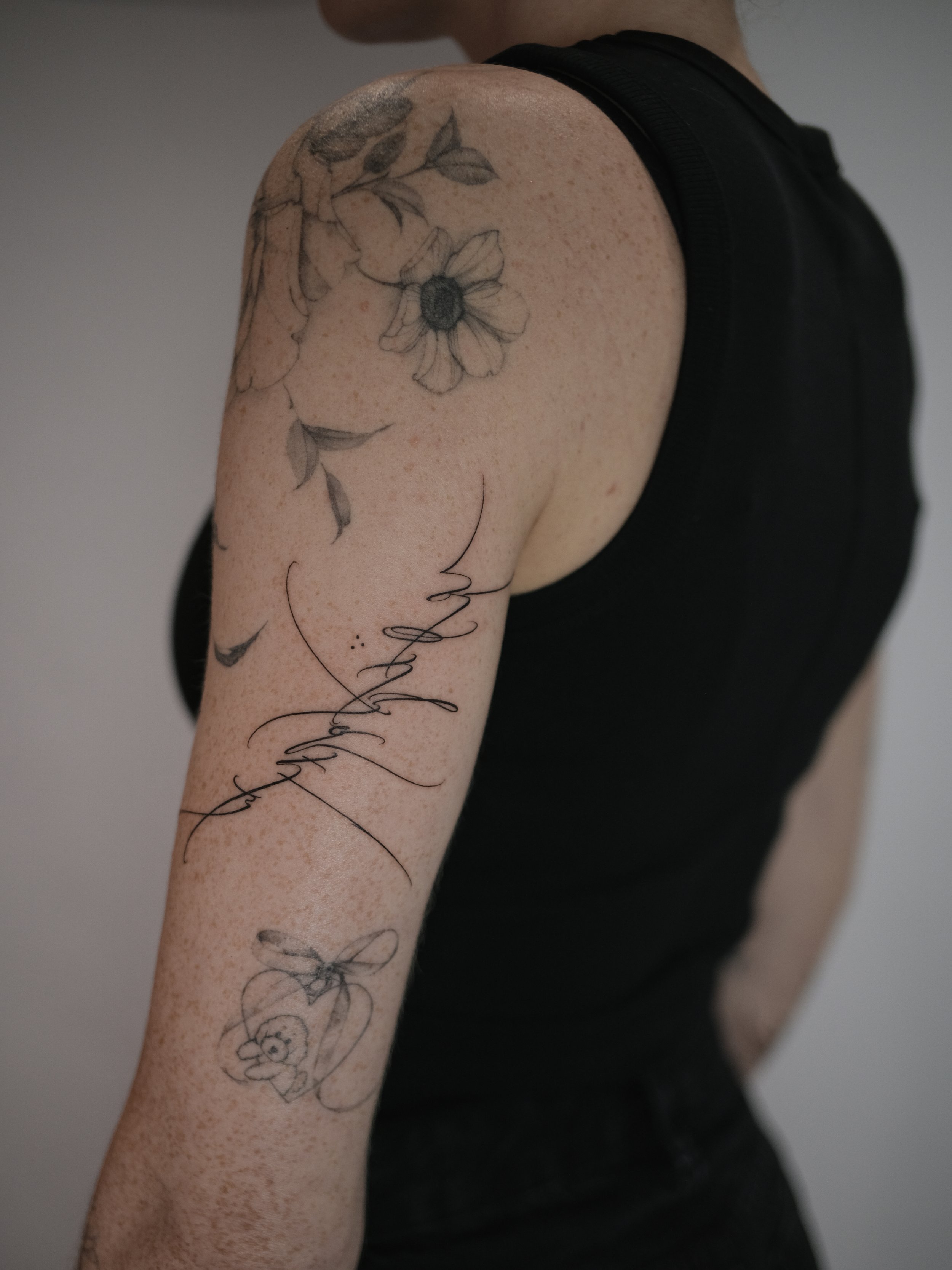 A person's upper arm with black and gray floral tattoos, including a large flower, leaves, and cursive script, against a plain background.