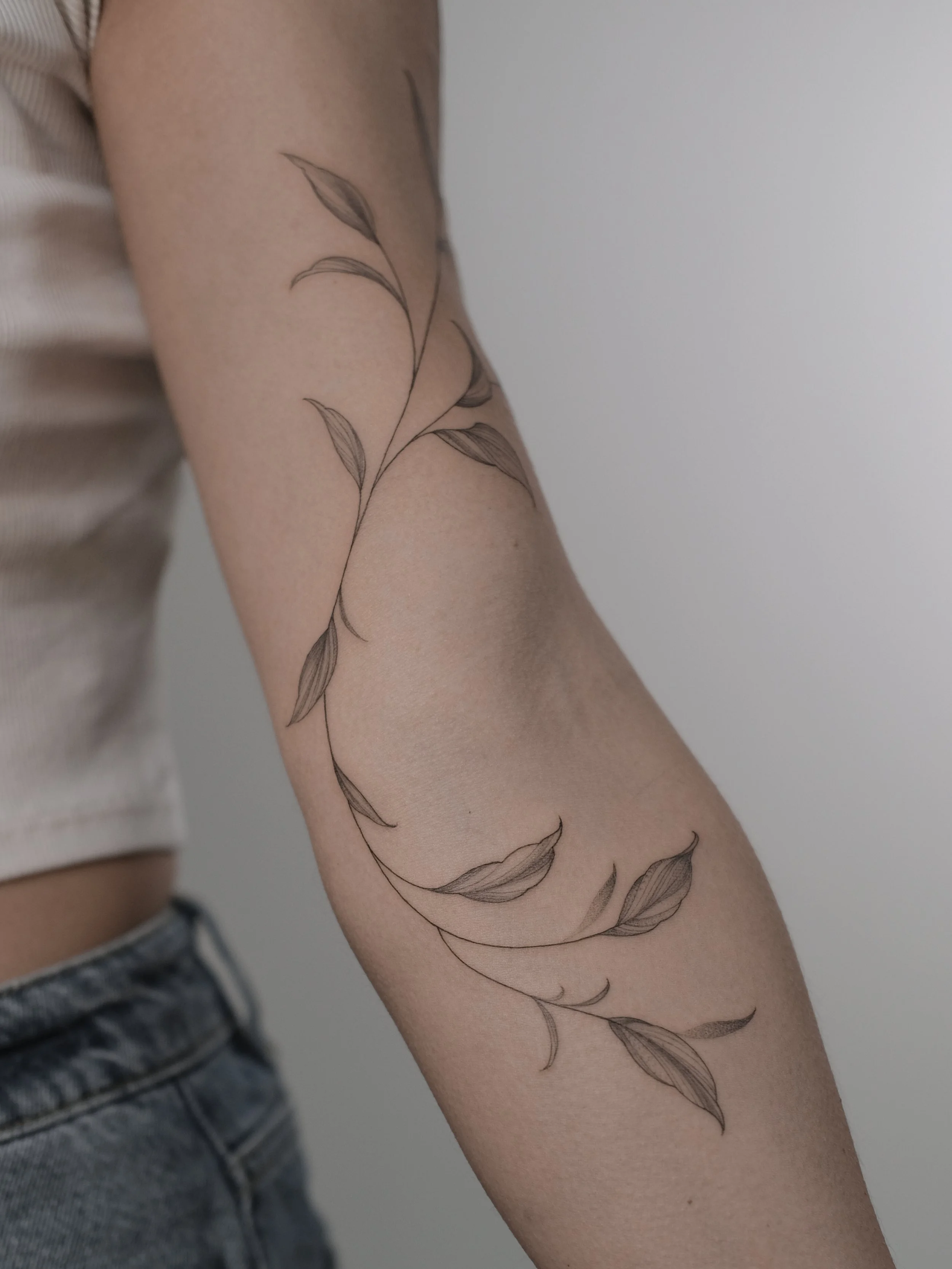 Close-up of a person's arm with a delicate black and gray botanical tattoo of a sprig with leaves.