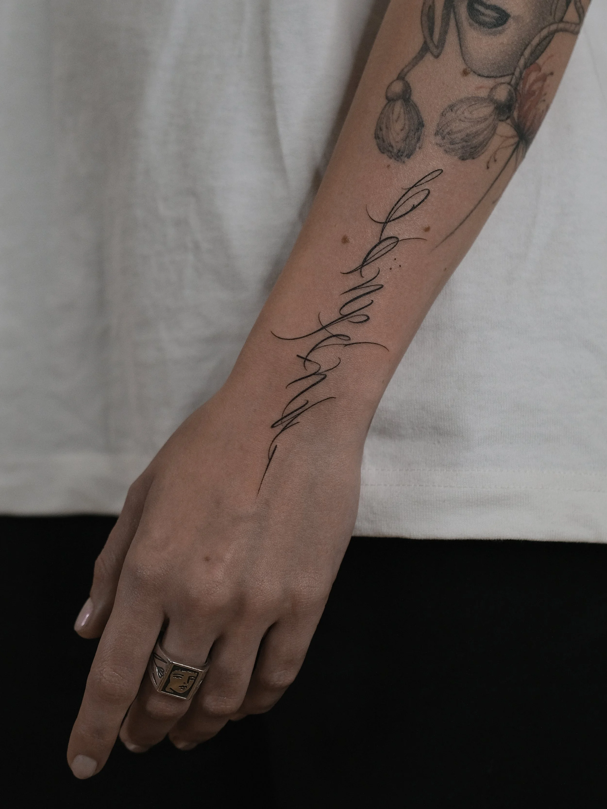 A person's hand and forearm with a tattoo and a ring on the finger, resting on a black surface, with a white shirt in the background.