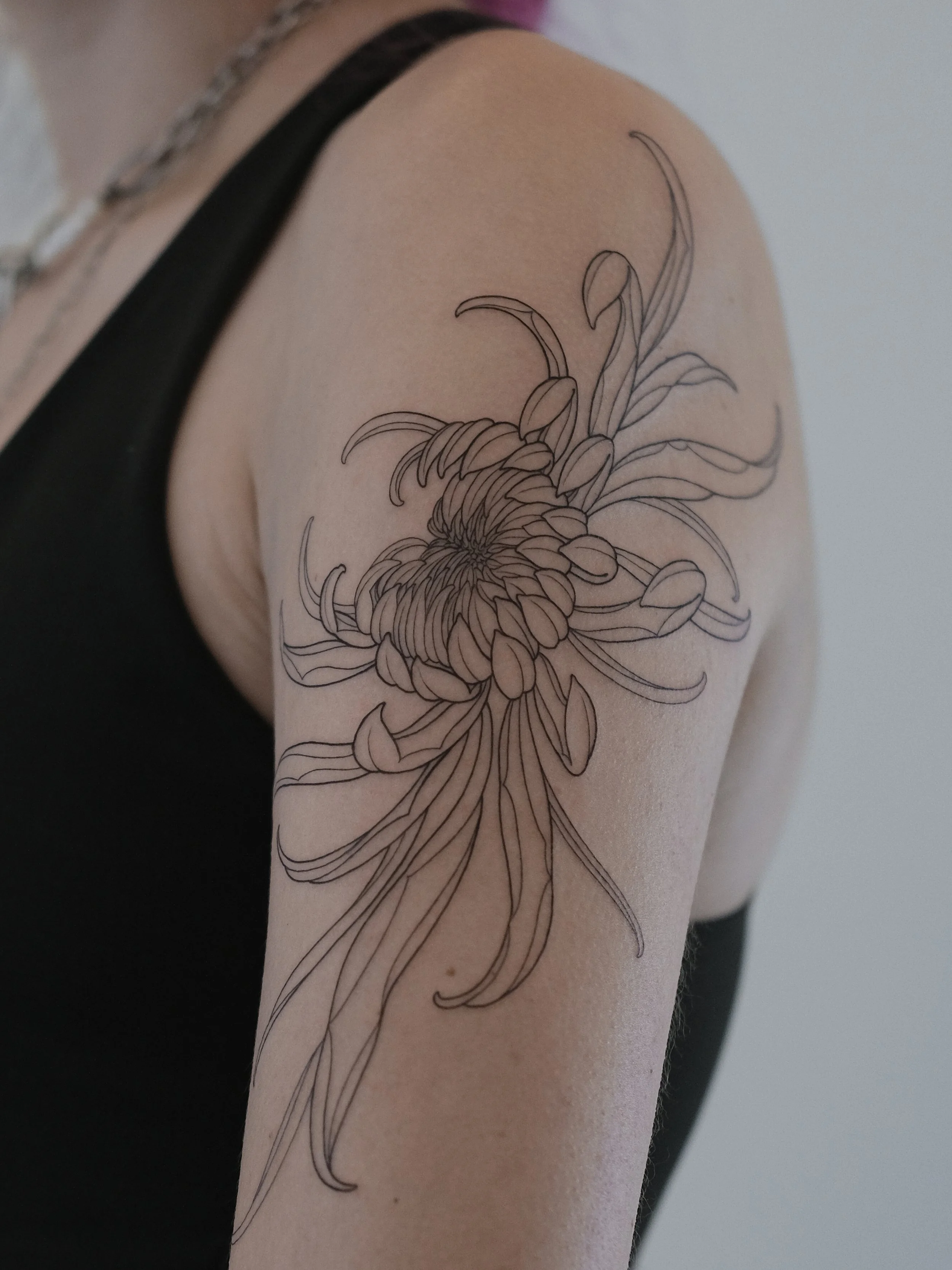 A person's arm with a detailed black line drawing tattoo of a large chrysanthemum flower and leaves.