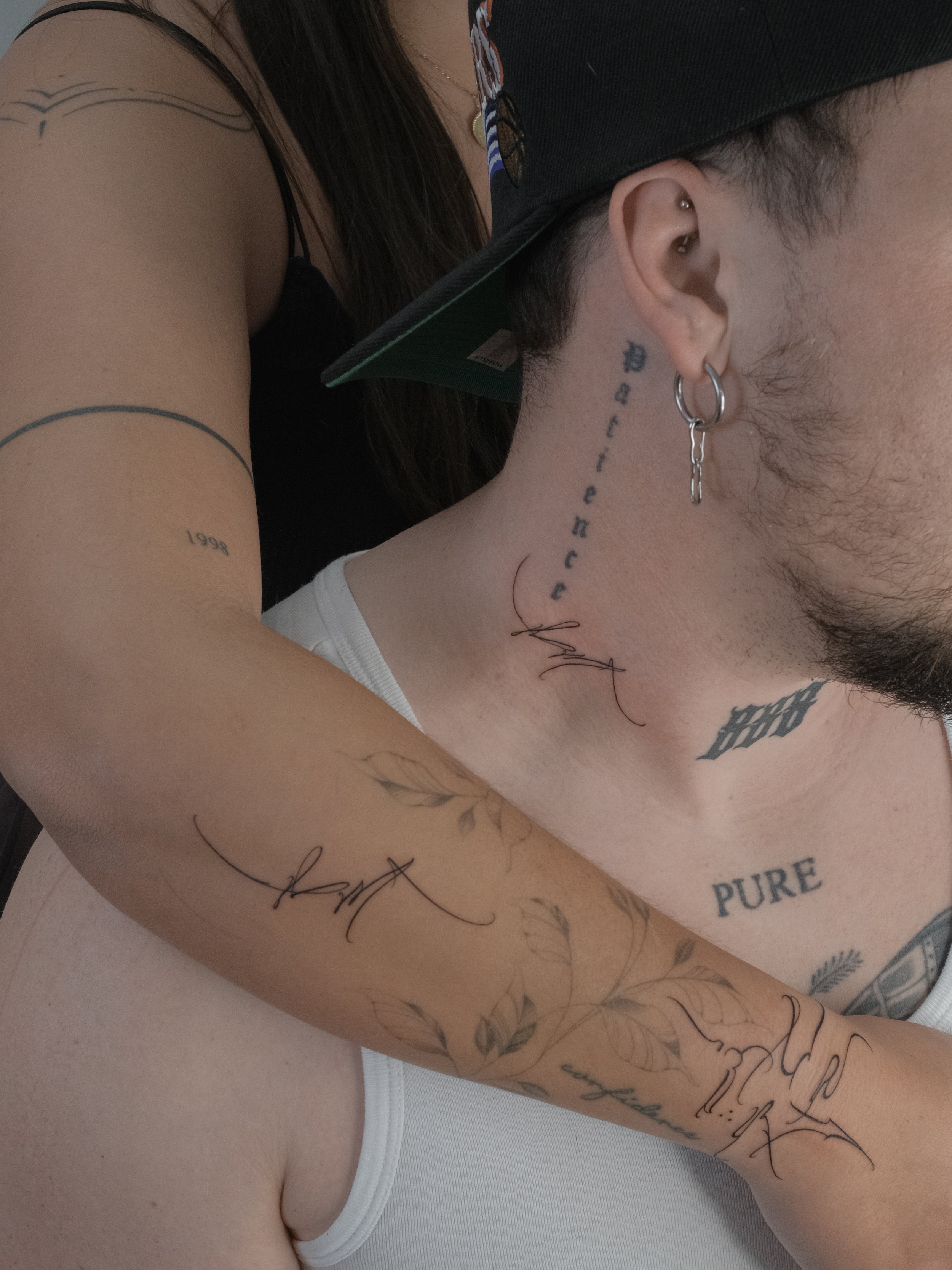 Close-up of a person's upper body showing various tattoos on their arm, neck, and chest, with some tattoos reading "1998," "PURE," and others in cursive and stylized fonts. The person is wearing a white sleeveless top, earrings, and a black cap.