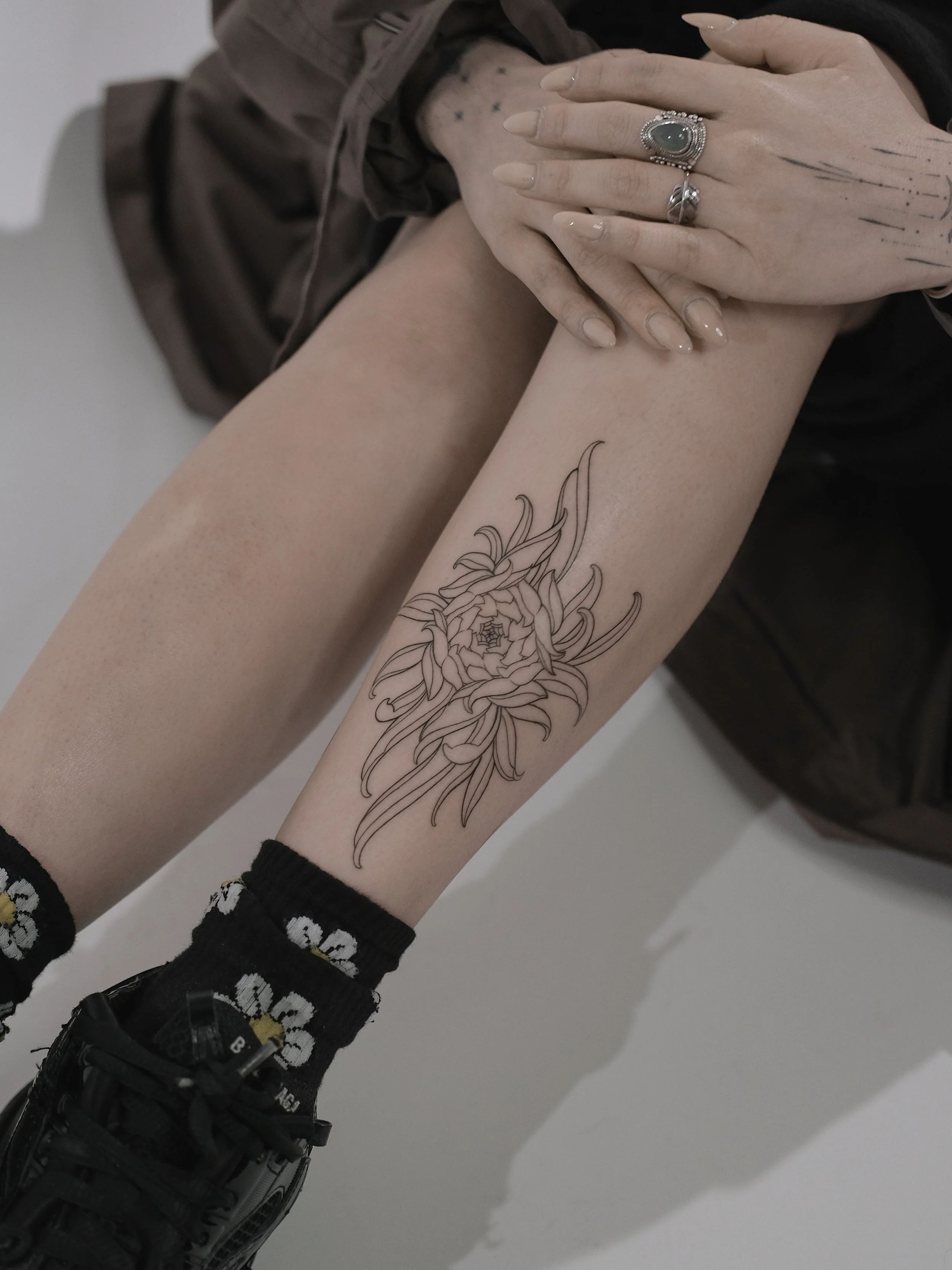 Person wearing black sneakers, black socks with a floral pattern, and denim shorts, revealing a tattoo of a floral design on their forearm. Person has multiple rings on fingers and clear nail polish. Hands are placed over their knee and thigh.