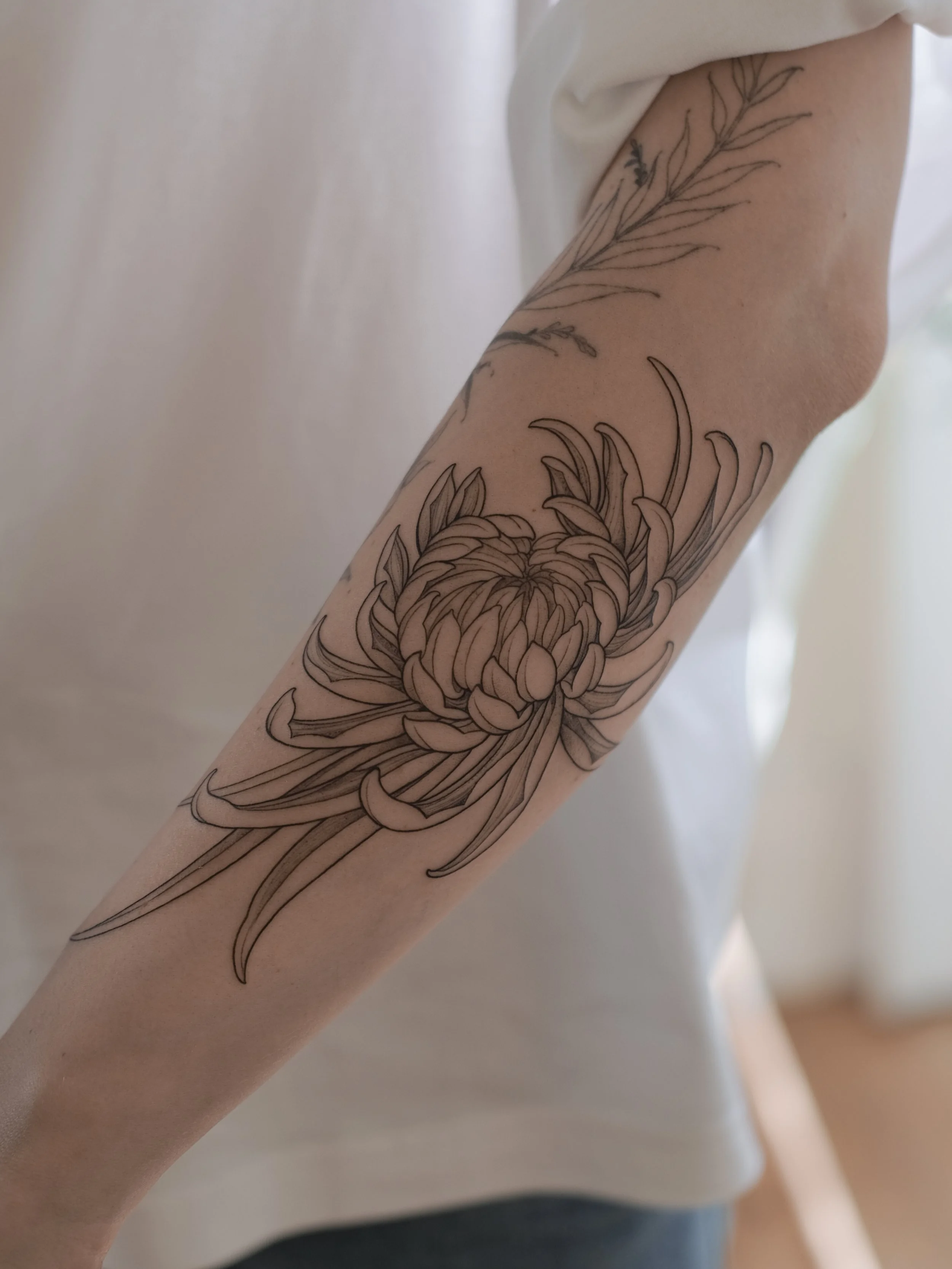 A person's arm with a black line tattoo of a detailed flower surrounded by leaves.