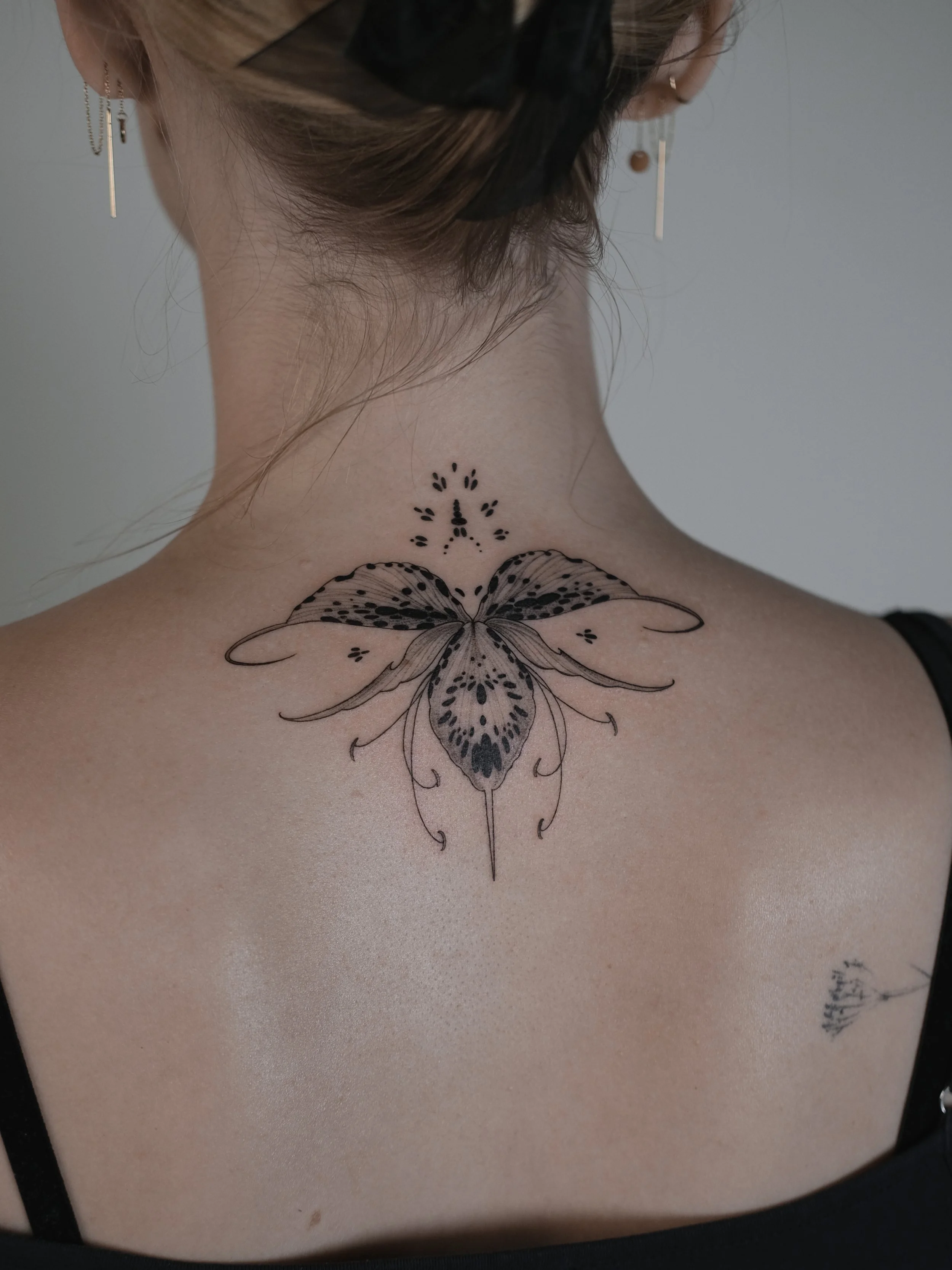 Back of a person's neck with a black bow in the hair and a large butterfly tattoo with floral details on the upper back, wearing earrings.