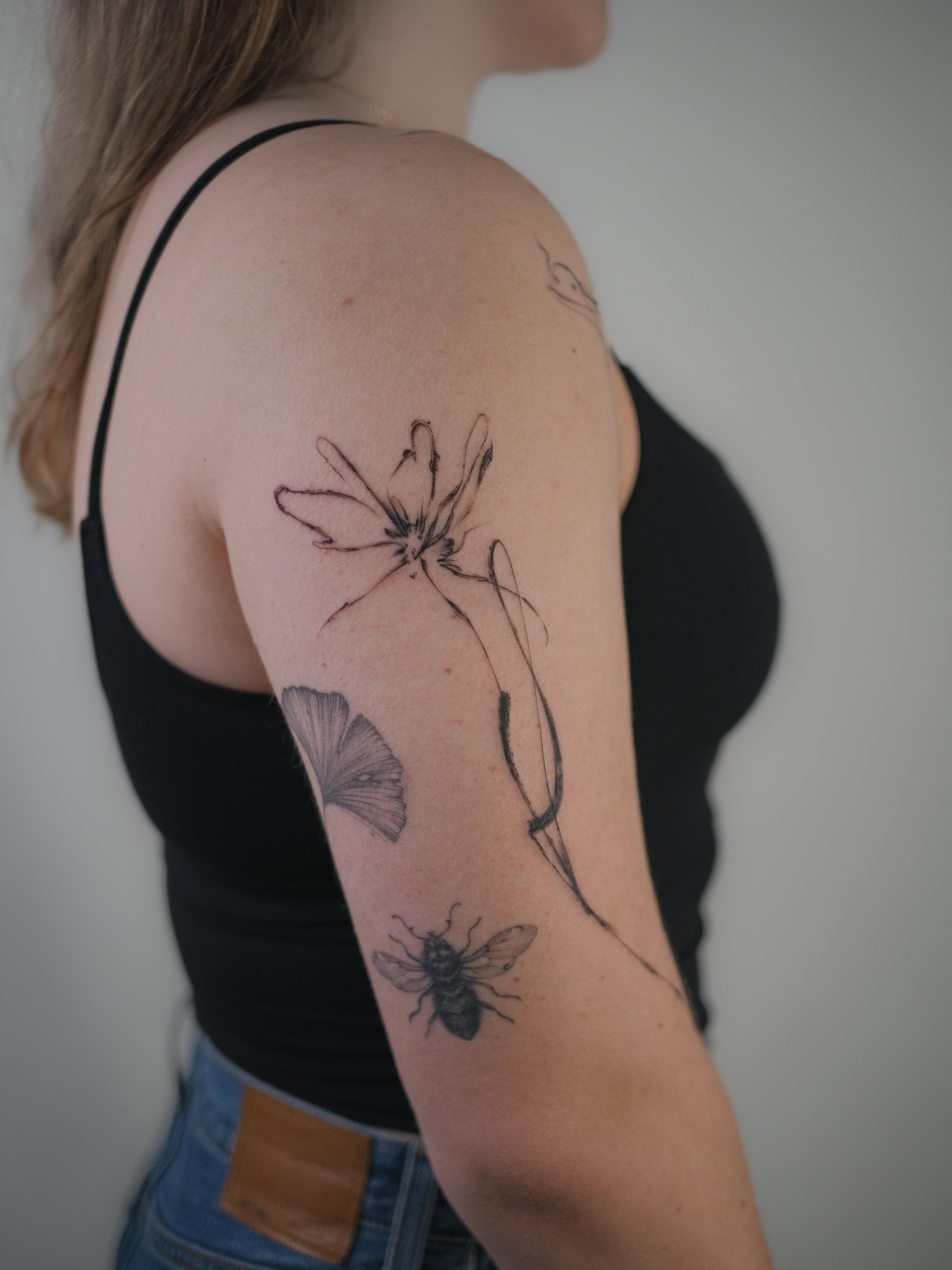 A woman's arm and shoulder with a tattoo of a line drawing butterfly, and part of her face and body visible. She is wearing a black spaghetti strap top and blue jeans.