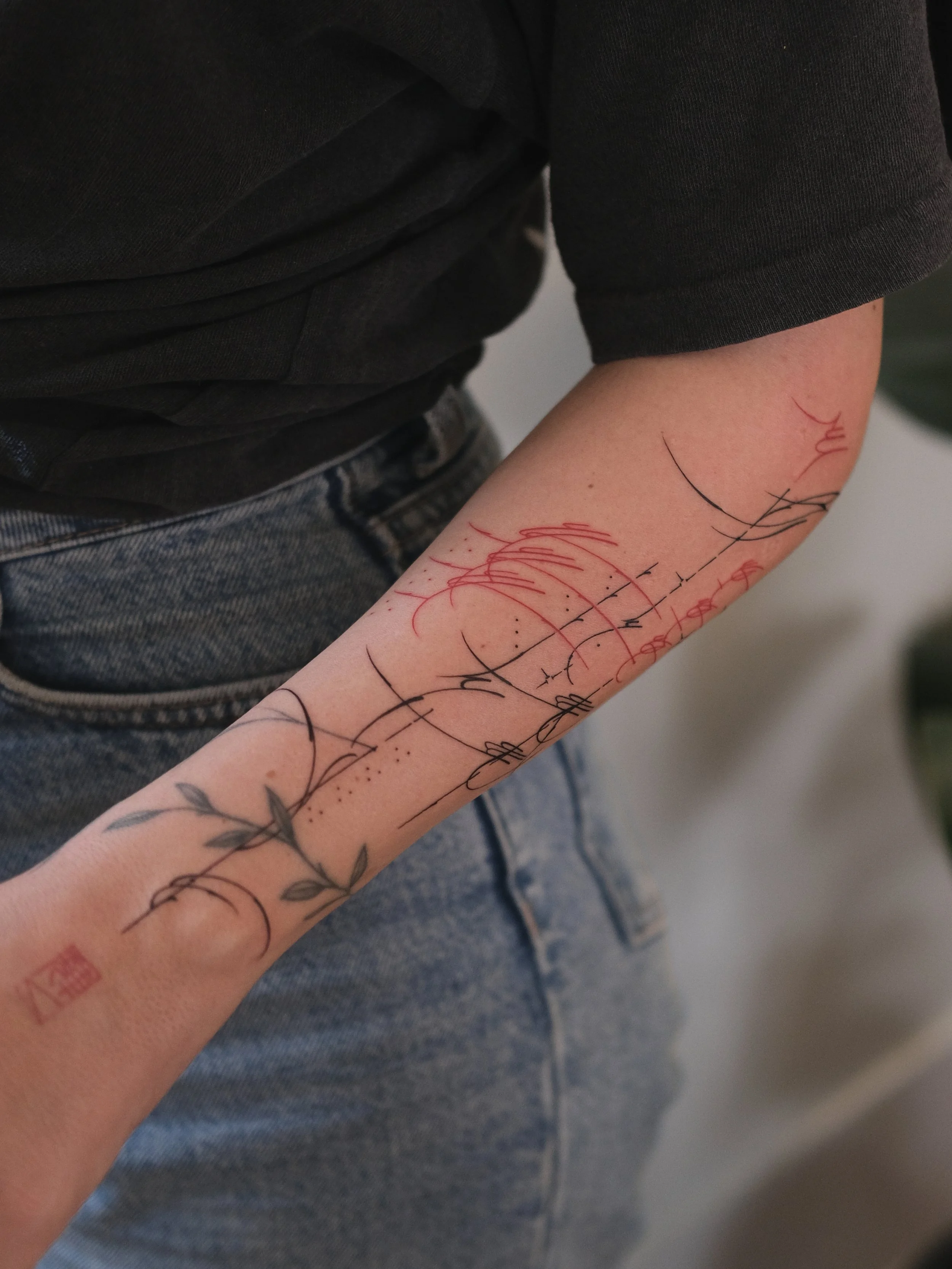 Close-up of a person's forearm with multiple tattoos, including floral and abstract designs, wearing a dark T-shirt and jeans.