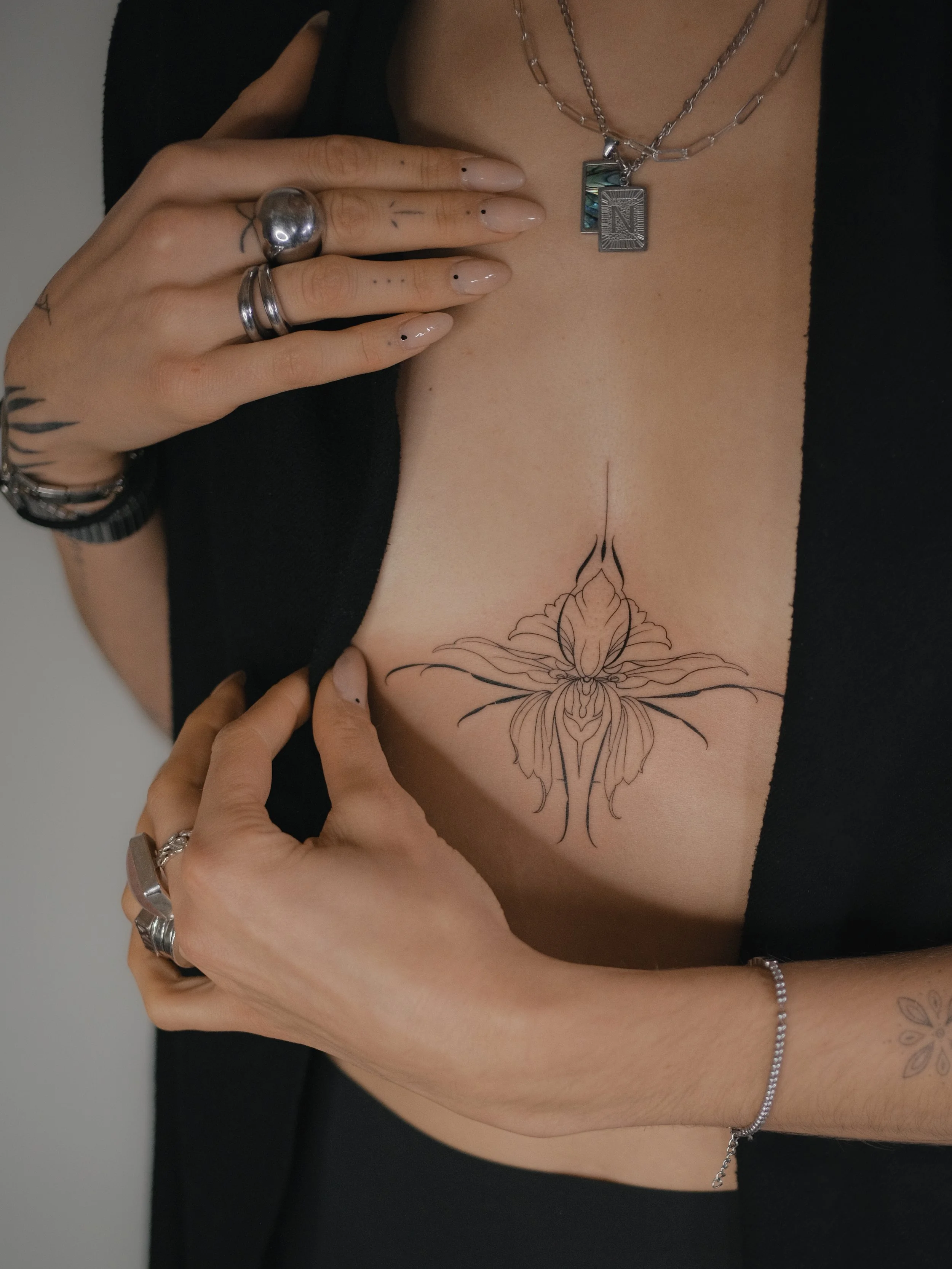 Close-up of a woman's chest showing a floral tattoo, jewelry including layered necklaces, rings, and bracelets, and hands with manicured nails displaying black dot nail art.