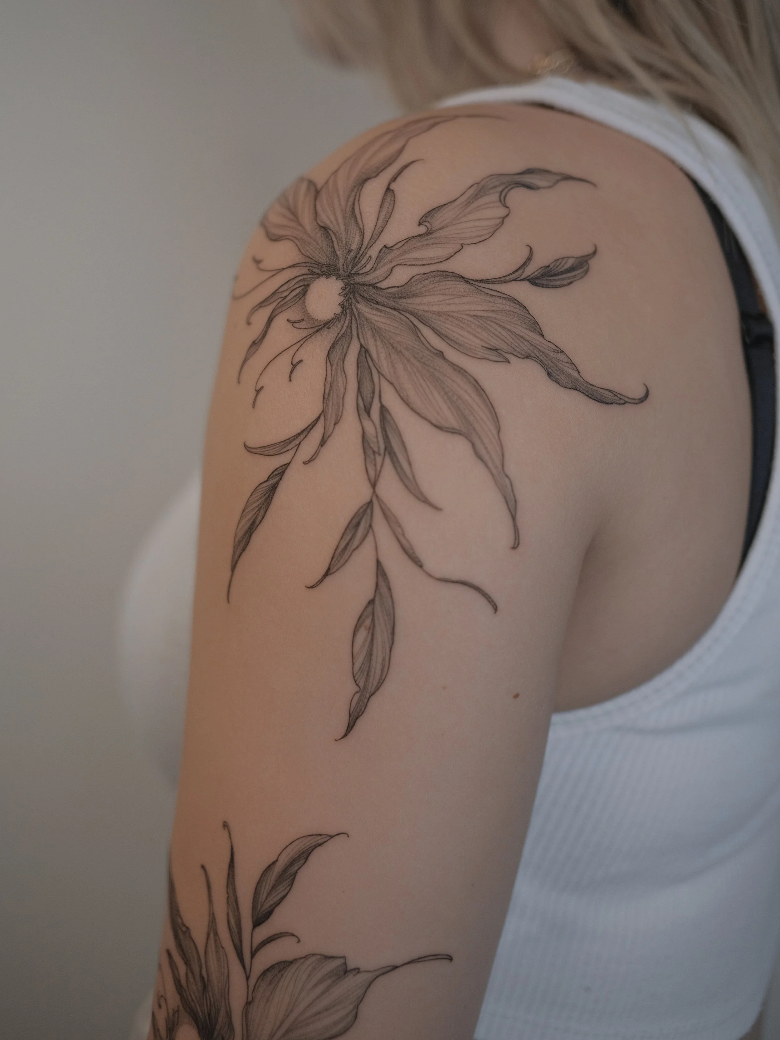 Close-up of a person's shoulder featuring a large, elegant floral tattoo in black ink with detailed petals and leaves.