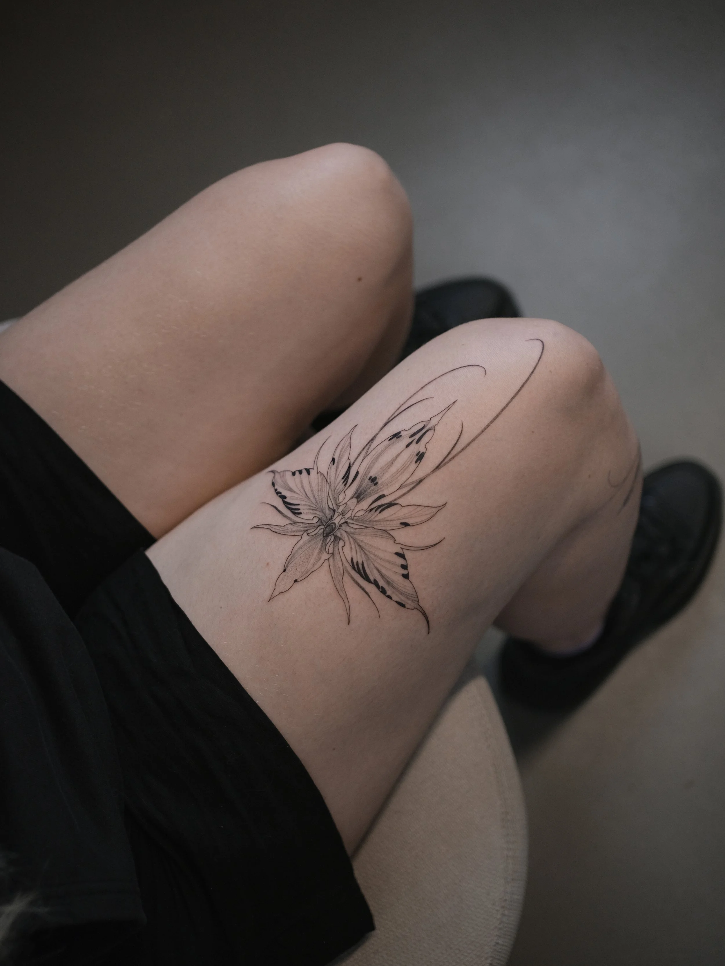 Person's thigh with a butterfly tattoo on the skin, some parts of black clothing and a grey background.