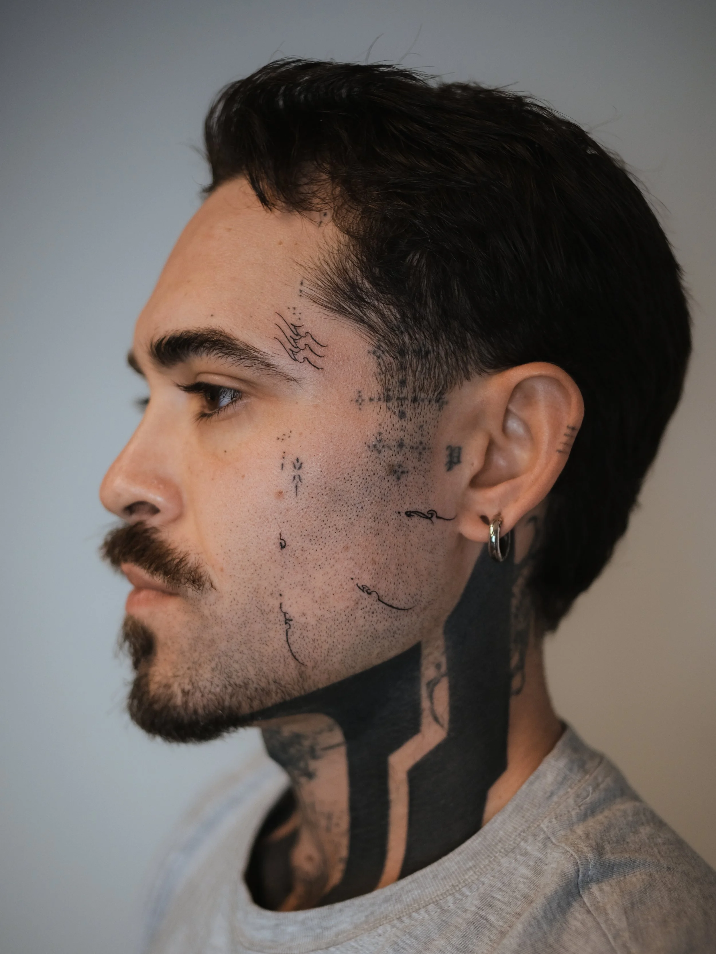 Close-up side profile of a young man with dark hair, tattoos on face and neck, and wearing a gray t-shirt.
