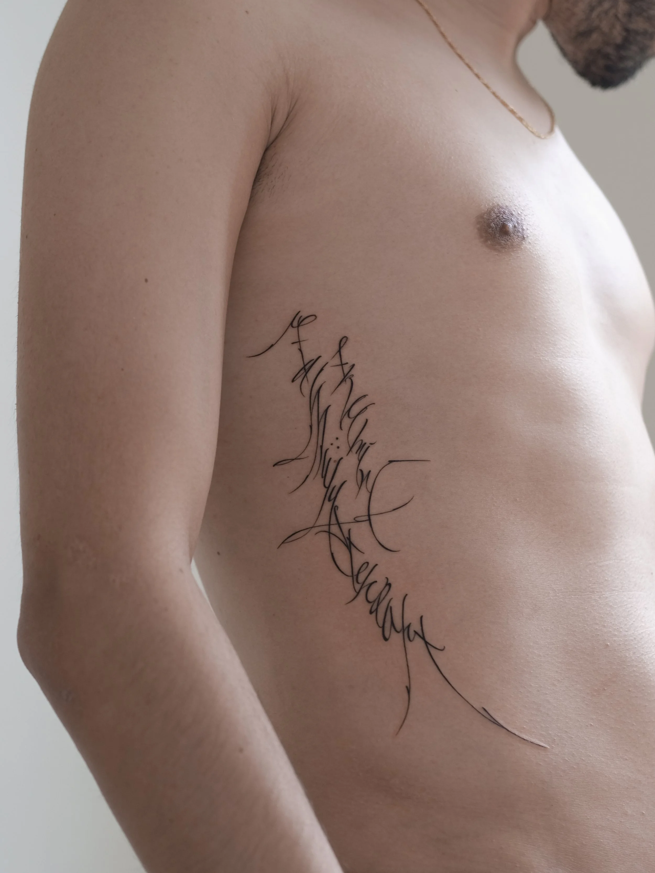 A close-up of a man's upper torso showing a tattoo written in cursive script on his side, with his face partially visible and a gold chain around his neck.