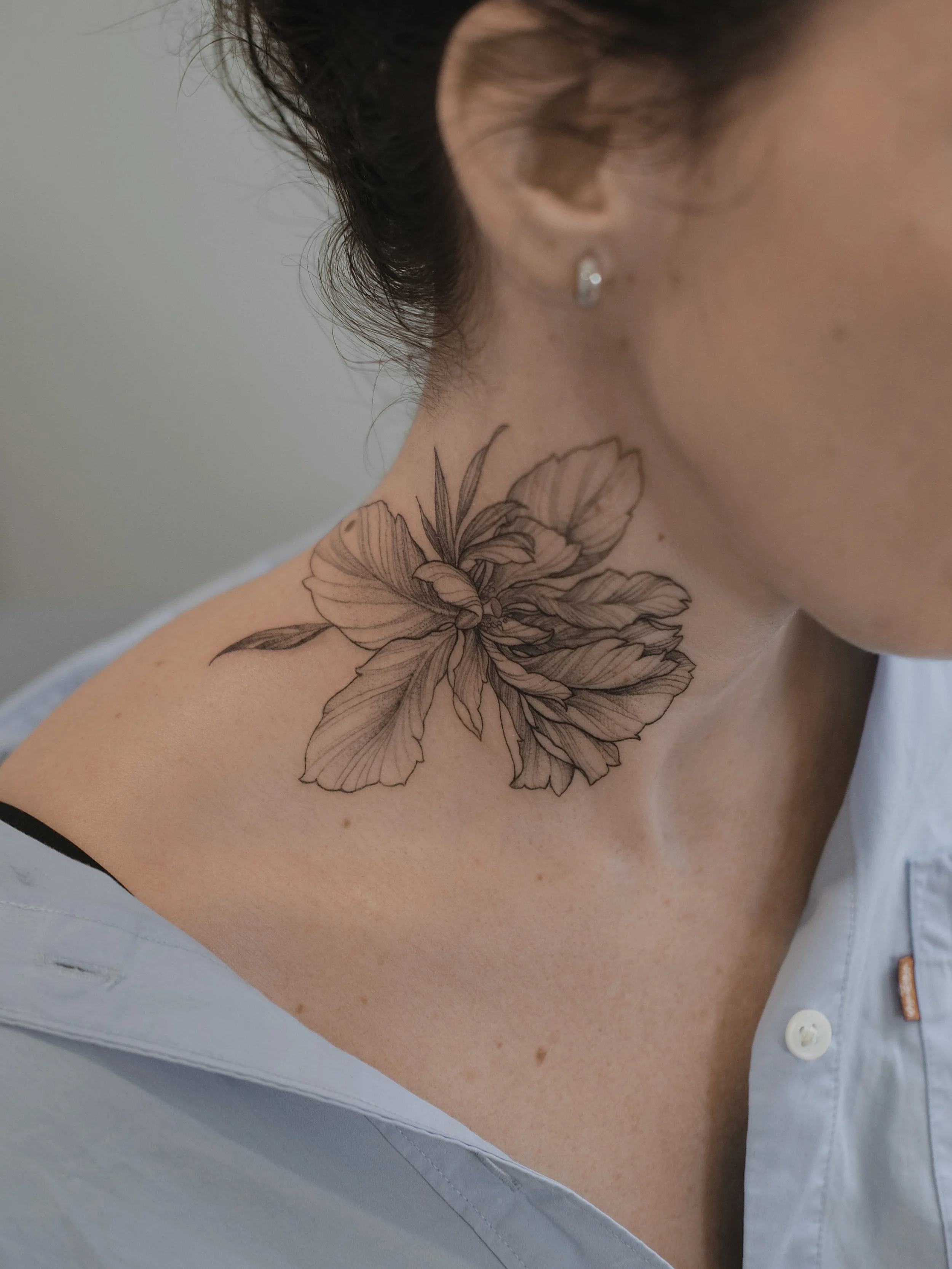 Close-up of a person's shoulder and neck with a floral tattoo, wearing a white shirt with buttons.