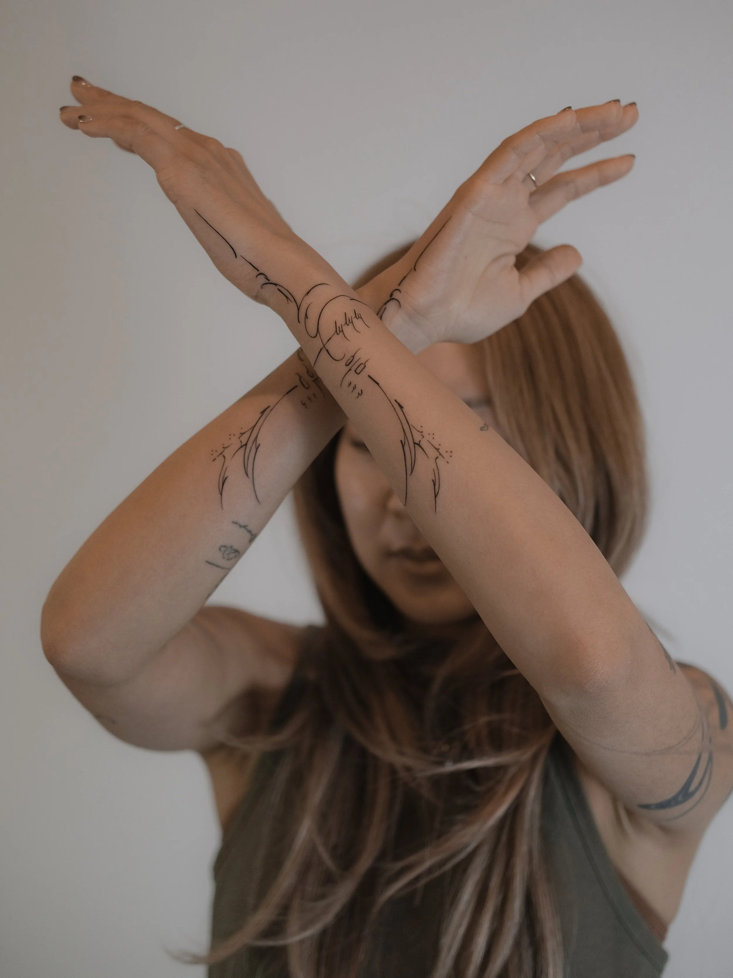 A woman with long brown hair and tattoos on her arms is crossing her hands in front of her face, obscuring her facial features.