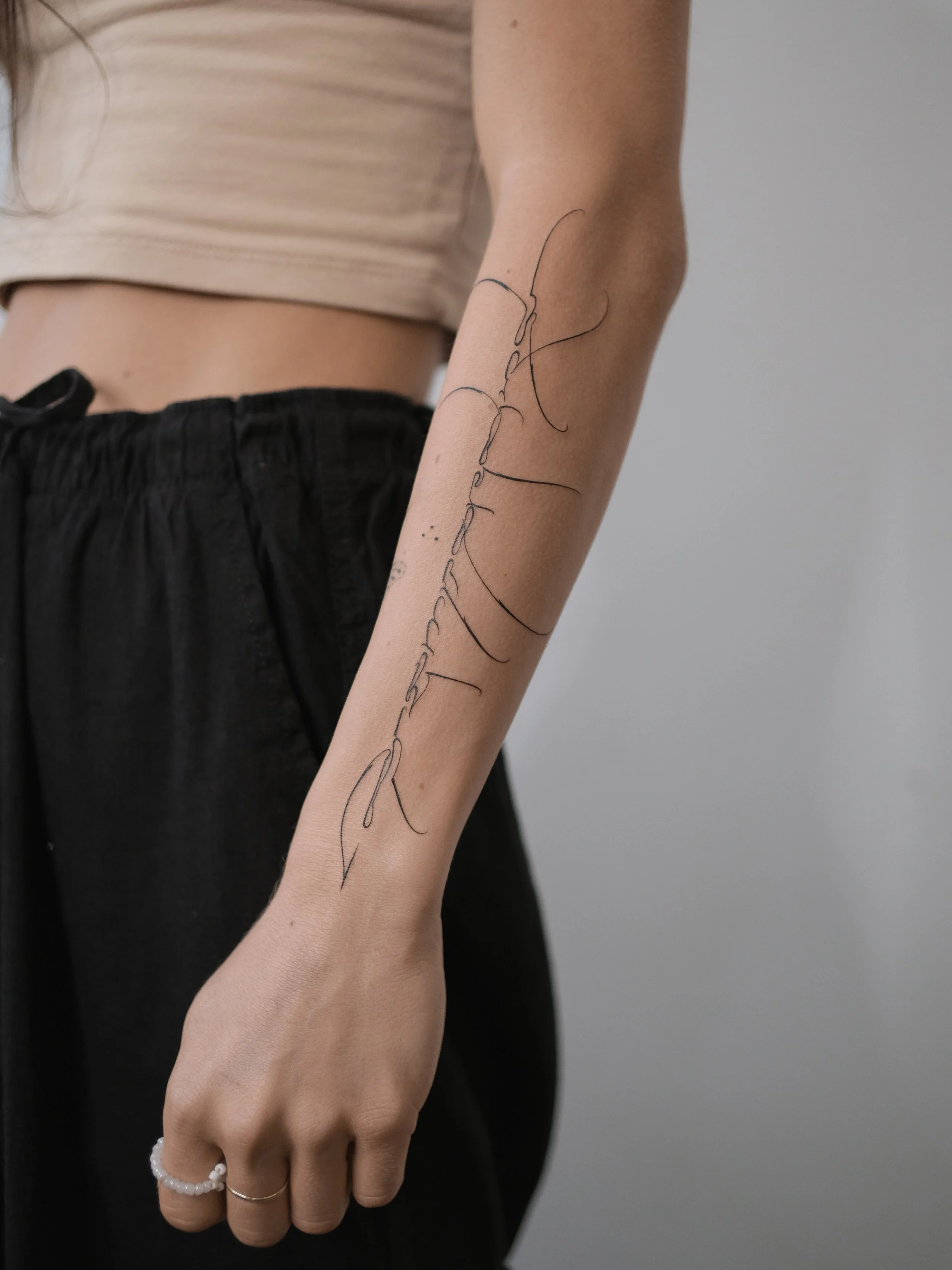 A woman's arm with a black tattoo of cursive text running along the outer forearm. The woman is wearing a beige top, black pants, a white beaded bracelet, and a silver ring on her ring finger. The background is a plain, light gray wall.