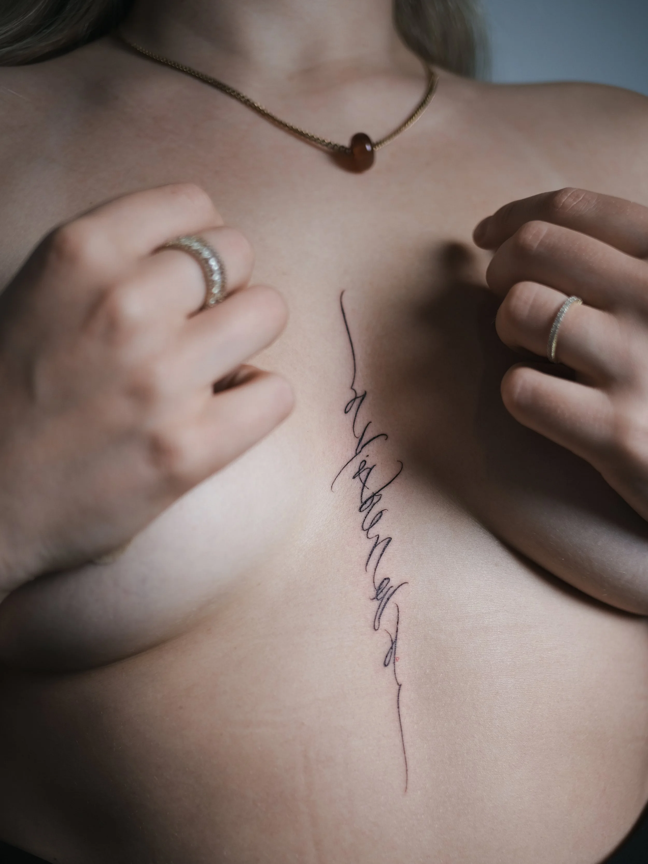 Close-up of a woman's chest with a tattoo of cursive writing along the center, hands with rings touching the skin, wearing a gold necklace with a dark pendant.
