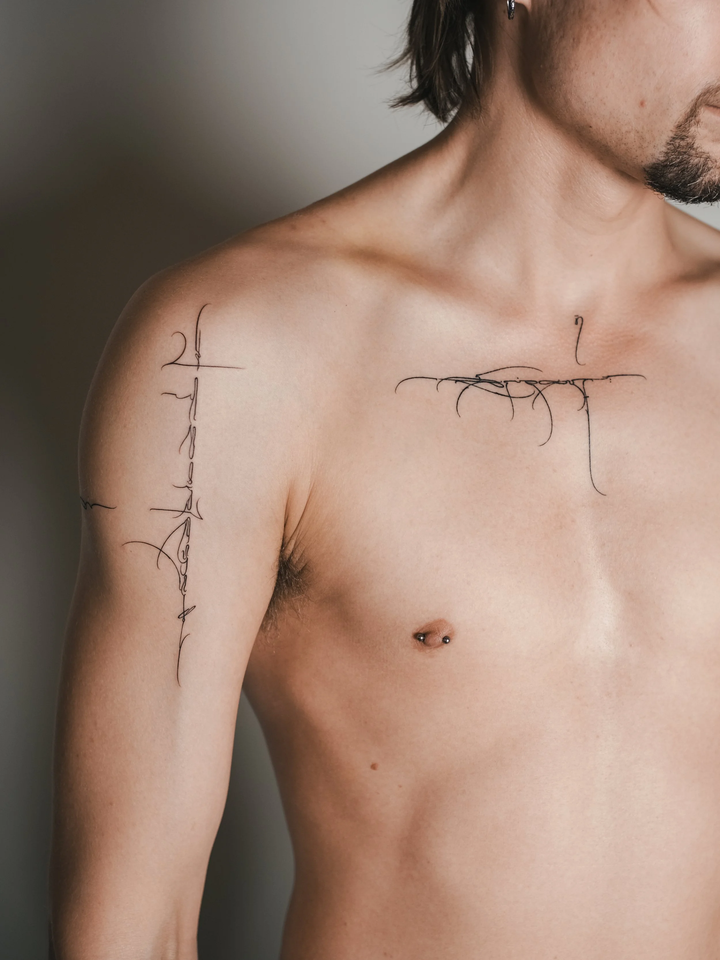 A close-up of a person's upper body, showing their shoulder, neck, and part of their face. They have tattoos on their shoulder and chest, and a belly button piercing. The person's skin is smooth and the background is plain and out of focus.