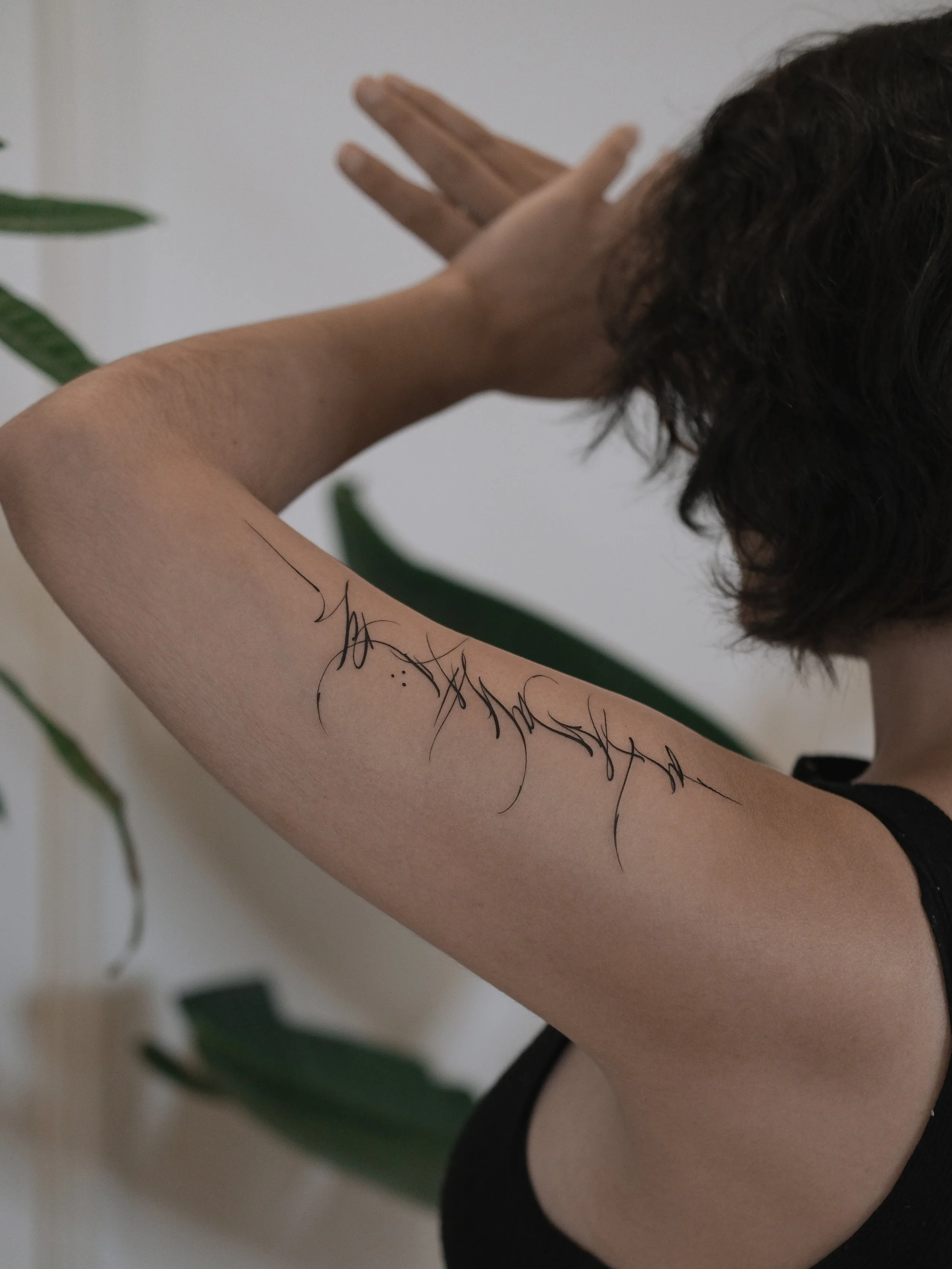 A person with dark, curly hair and light skin is resting their head against their bent arm, which has a tattoo of a curvy line and dots. The person is wearing a black sleeveless top, and there is a plant with broad green leaves in the background.