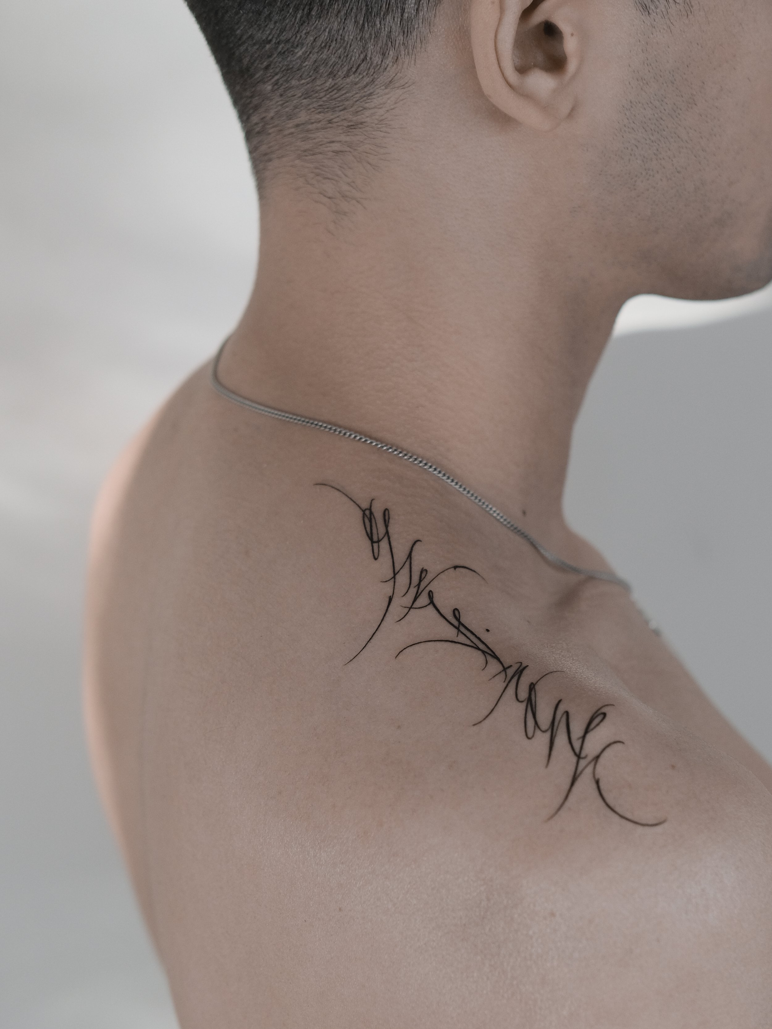 Close-up of a person's neck and shoulder, showing a tattoo with cursive writing and a silver necklace.