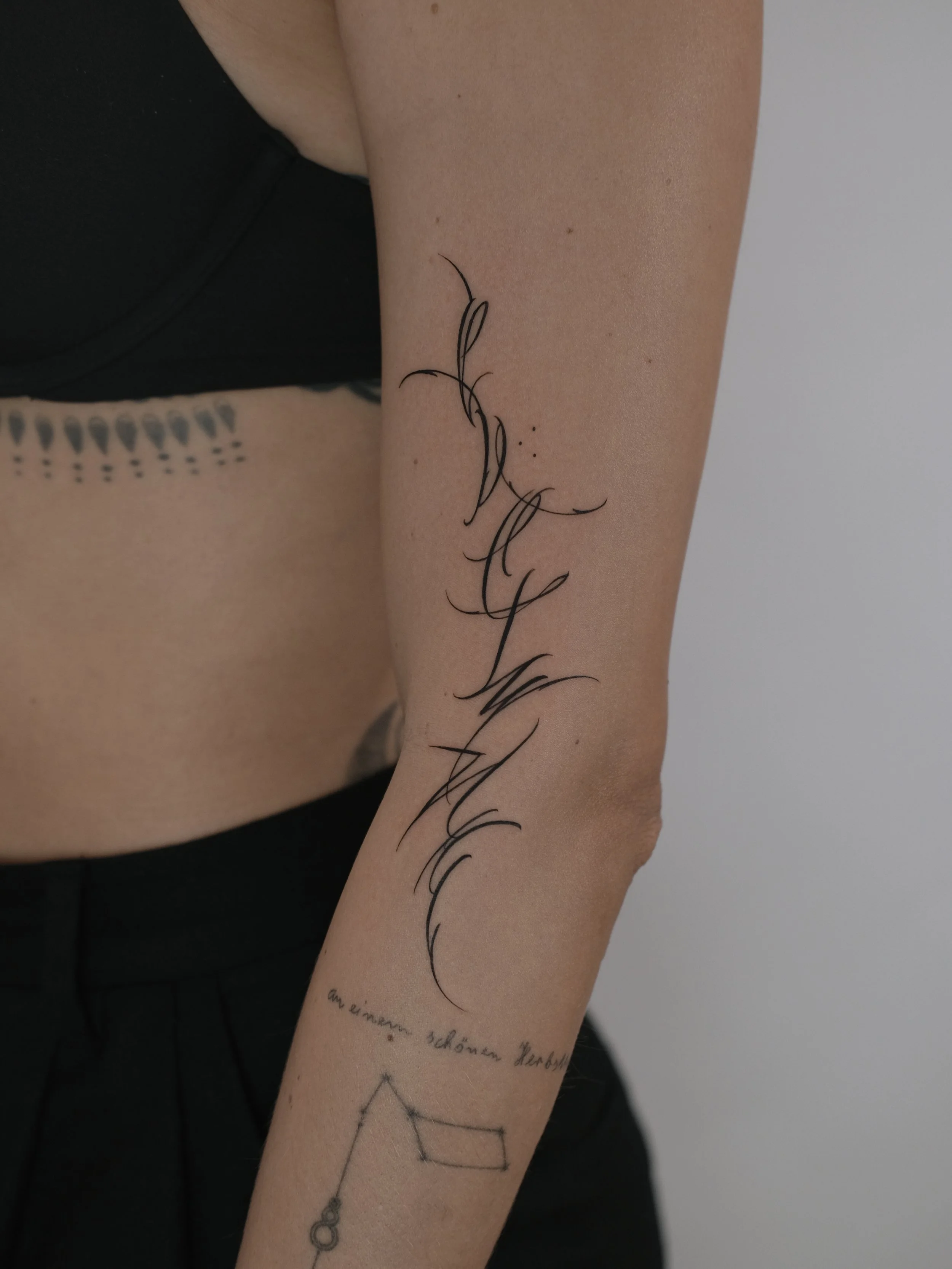 Close-up of a person's arm with a black ink tattoo of a stylized, flowing line design and German text beneath it. Part of the person's black clothing is visible.