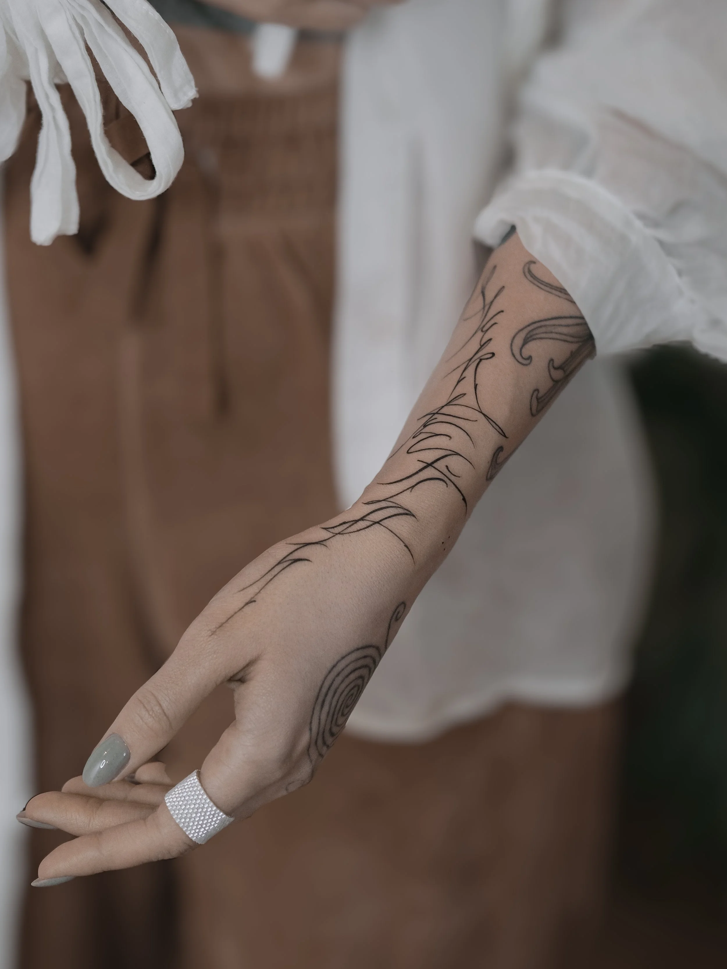 Close-up of a person's arm with black tattoo lines and a silver-colored ring on the index finger, wearing a white long-sleeve shirt, with a wooden dresser or cabinet in the background.
