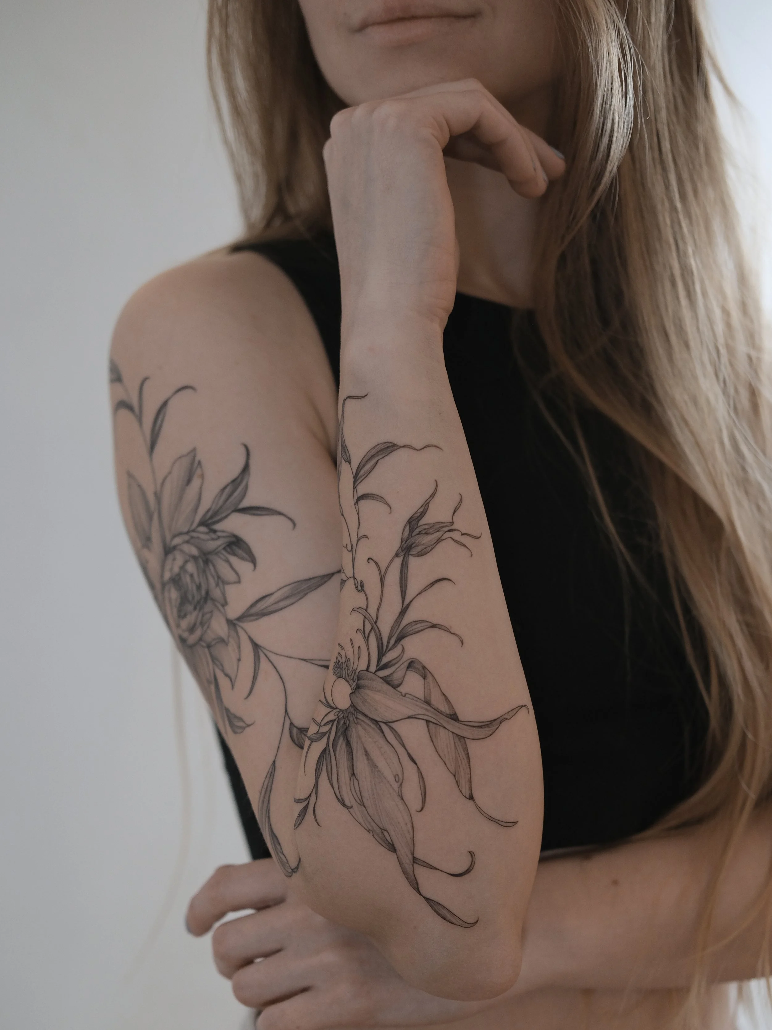 Close-up of a woman's arm with a large floral tattoo and part of her face, with her hand resting near her chin, wearing a black sleeveless top.