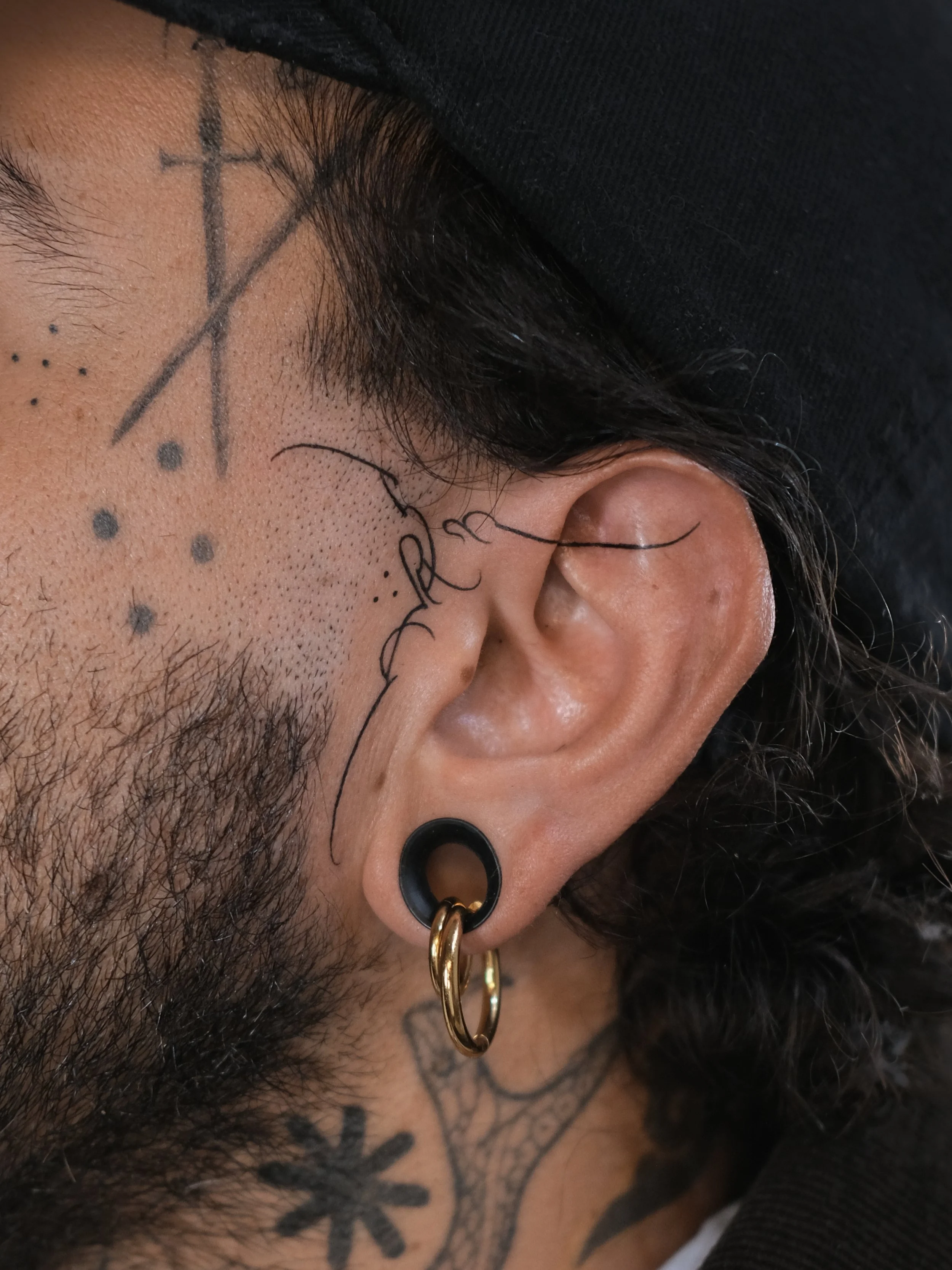 Close-up of a person's face with tattoos near the eye, including a cross and dots. The person has multiple earrings, including black earrings and gold hoops, and is wearing a black cap or hoodie.
